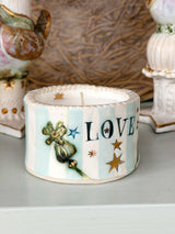A small decorative ceramic candle holder with a white and light blue striped pattern, featuring the word 'LOVE' in dark blue letters, gold stars, and a green fairy illustration. The candle is shown on a tabletop with other vintage-style ornaments blurred in the background.