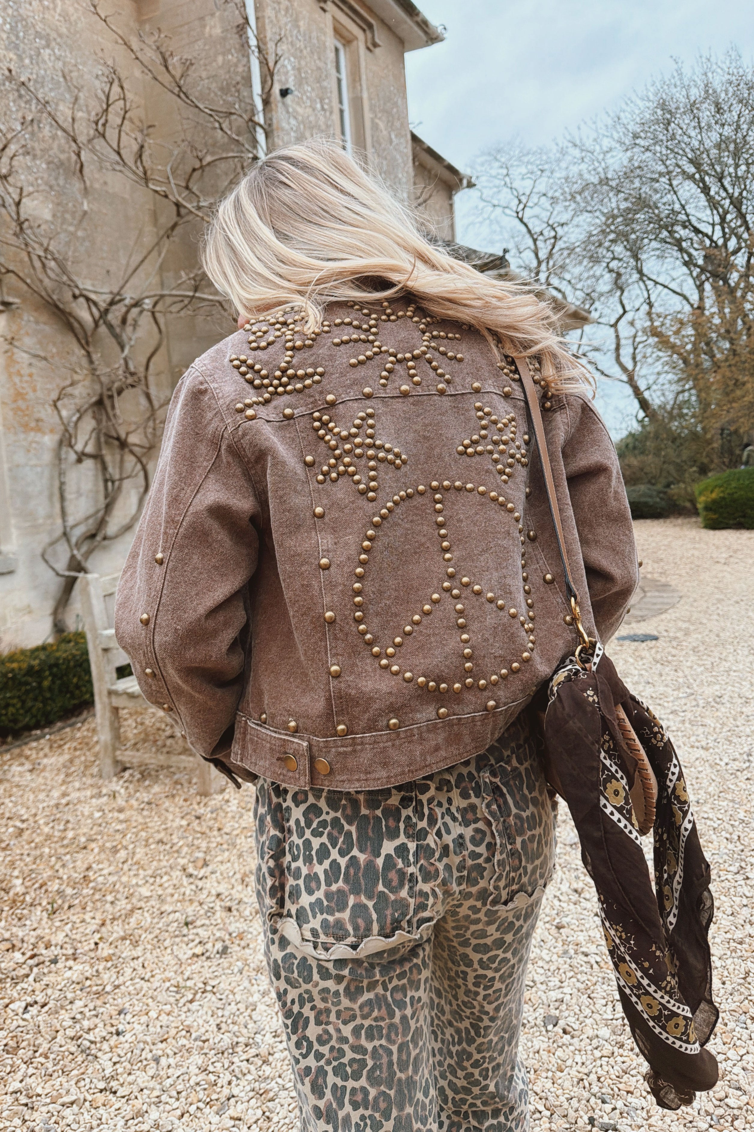 Woman wearing a brown jacket with brass stud peace sign and heart designs on the back, paired with leopard print pants outdoors.