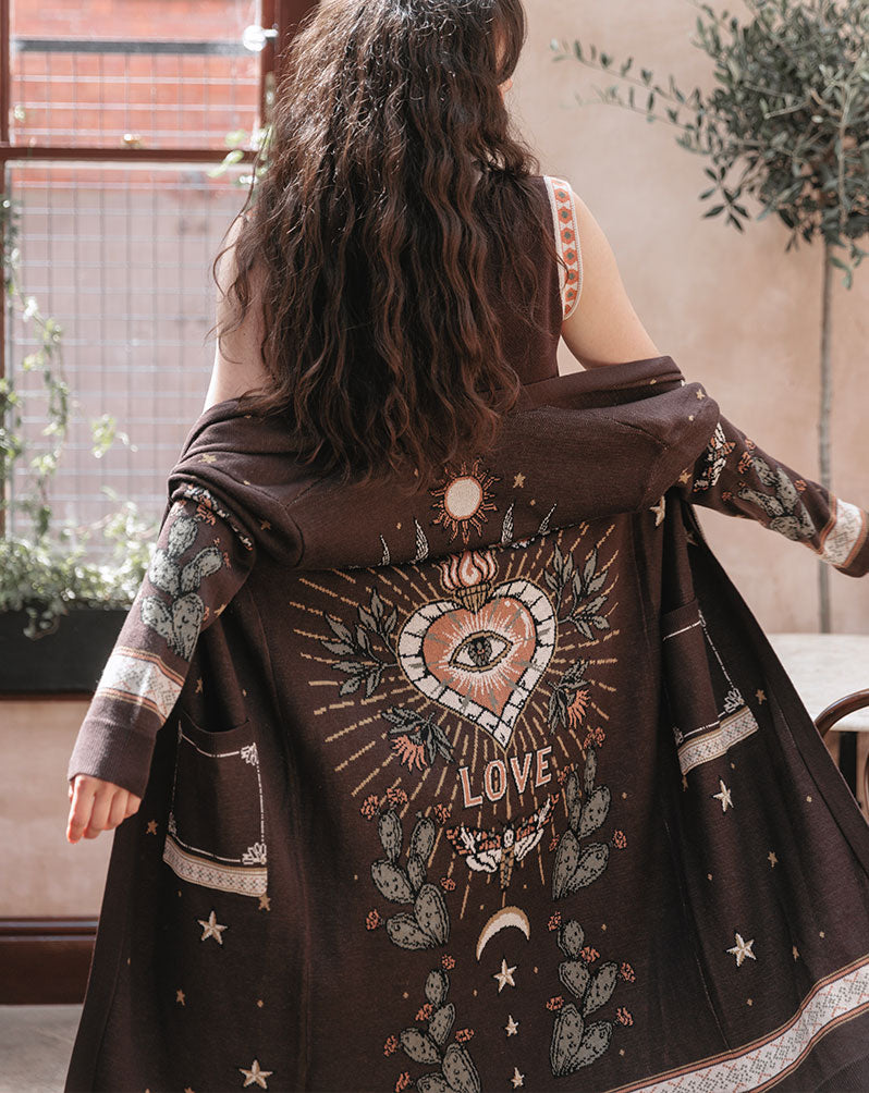Person wearing a dark brown knit cardigan with a celestial western-inspired knit design featuring a sacred heart, sun, moon, cacti, stars, and the word "LOVE" on the back.