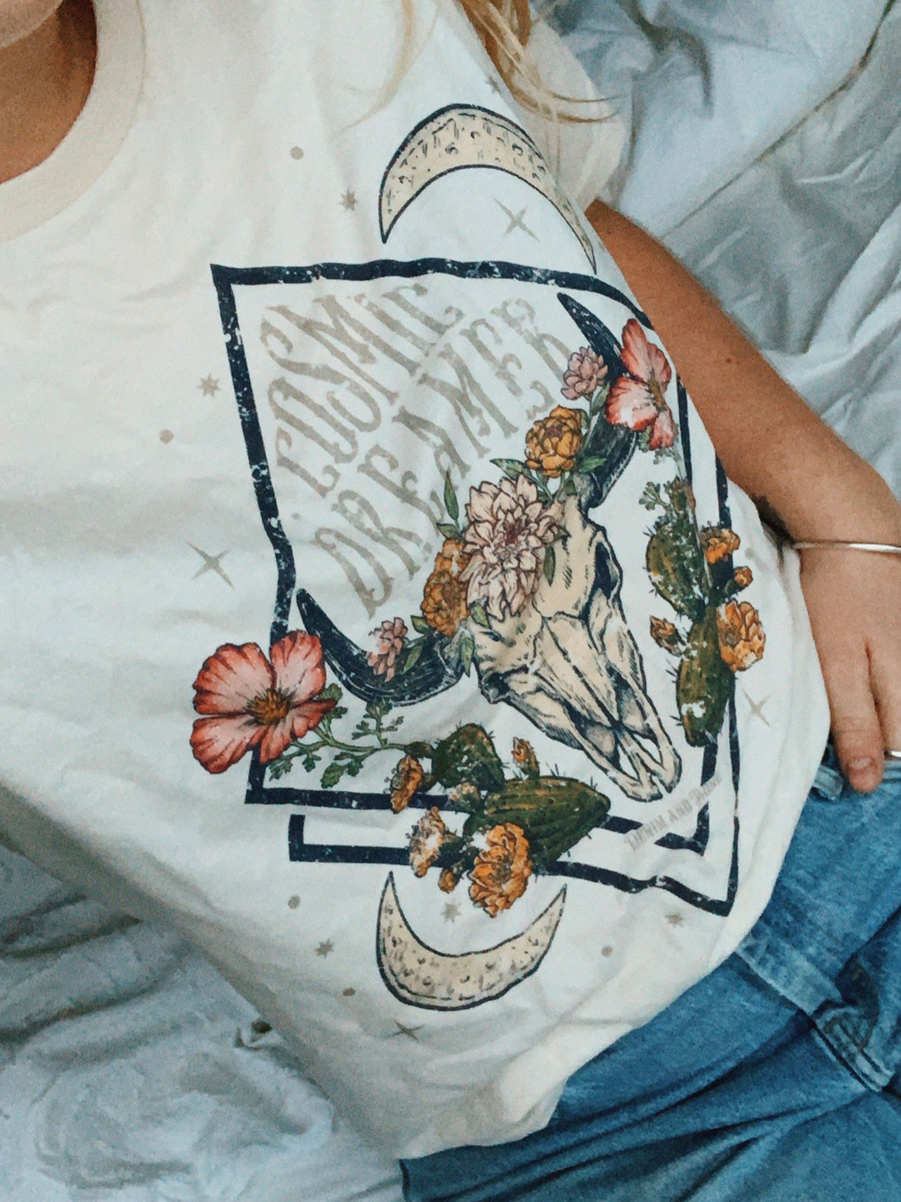 Close-up of a white graphic T-shirt featuring a floral longhorn skull design with crescent moons, desert cacti, and "Cosmic Dreamer" text.
