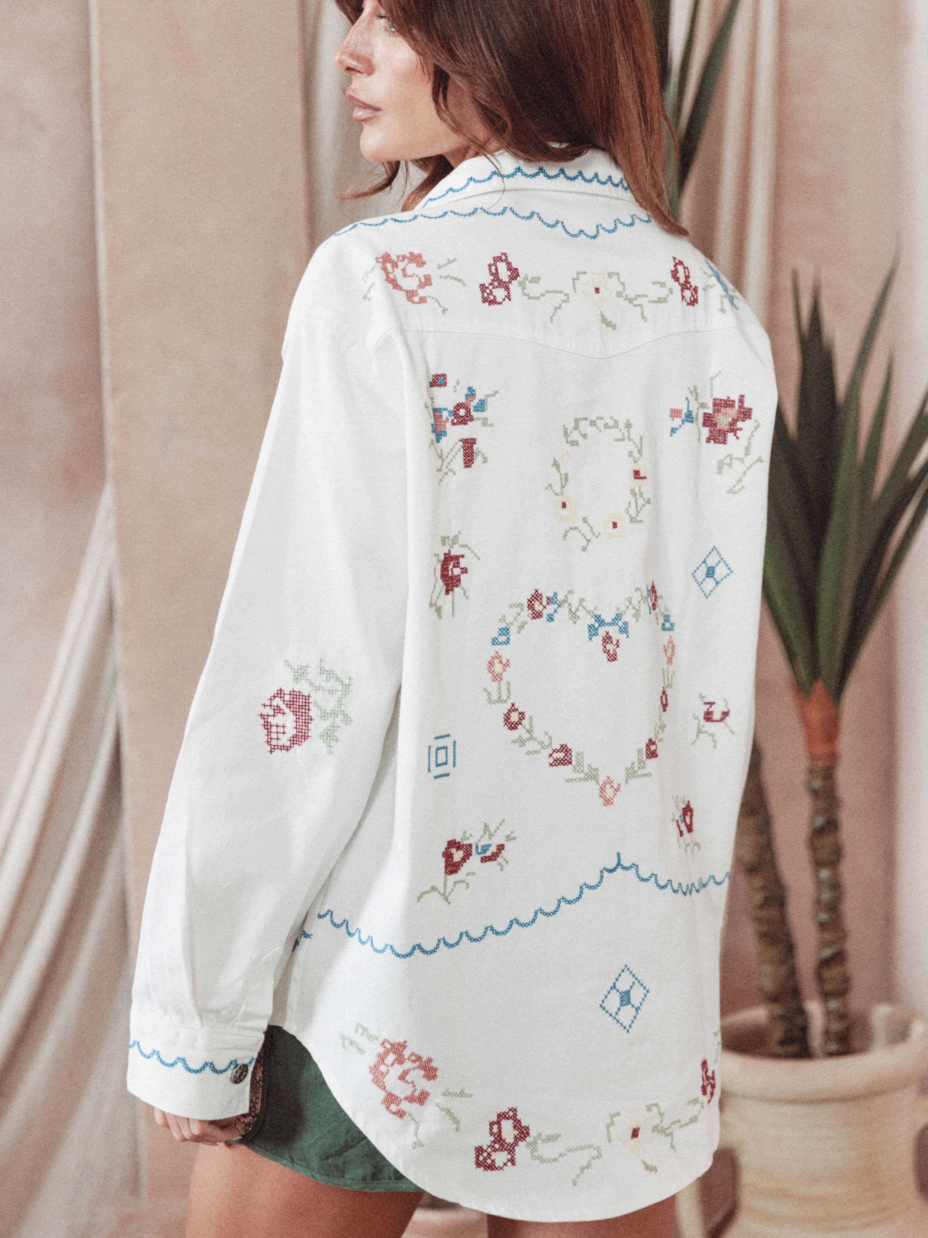 Back view of a person wearing a cream button-up shirt featuring colorful floral cross-stitch embroidery, heart motifs, and scalloped blue embroidered trim.