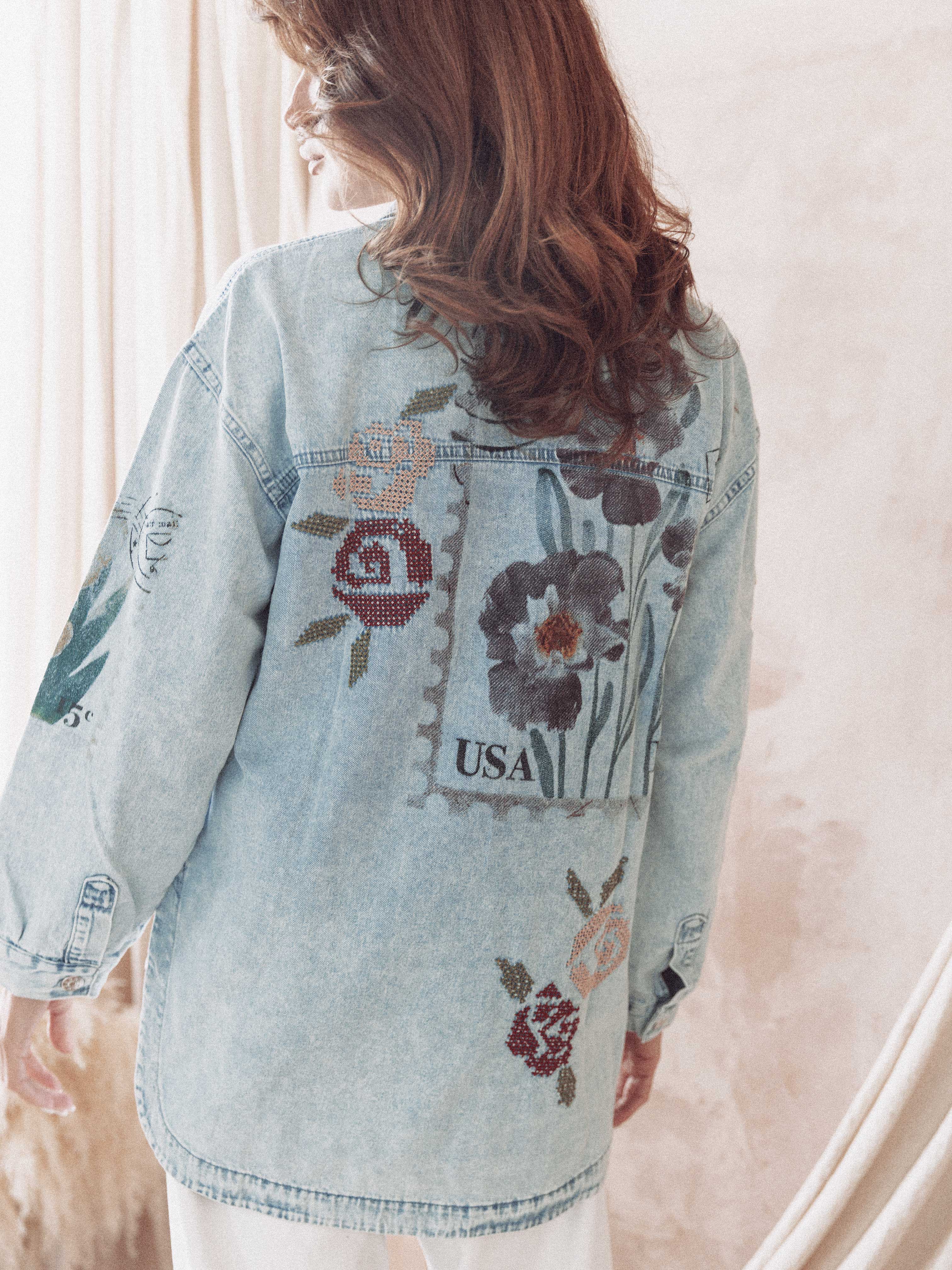 Back view of a person wearing a light wash denim button-up shirt featuring floral embroidery and vintage-inspired postage stamp patchwork with "USA" text.