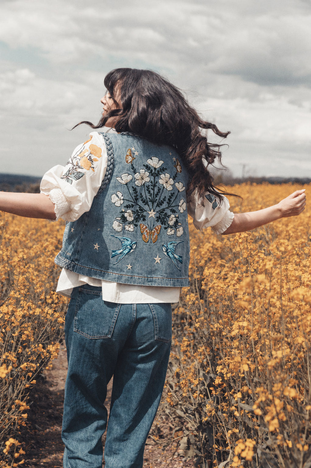 Person wearing a blue embroidered denim vest over a white embroidered blouse in a field of yellow wildflowers.