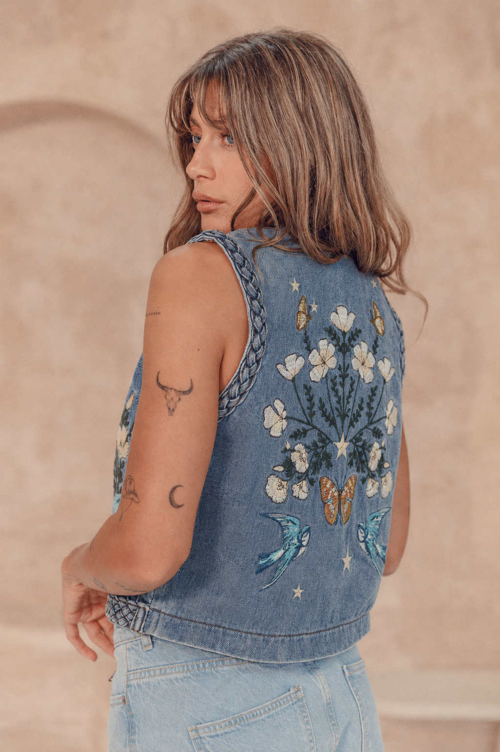 Woman wearing a denim vest with floral and butterfly embroidery against a beige background