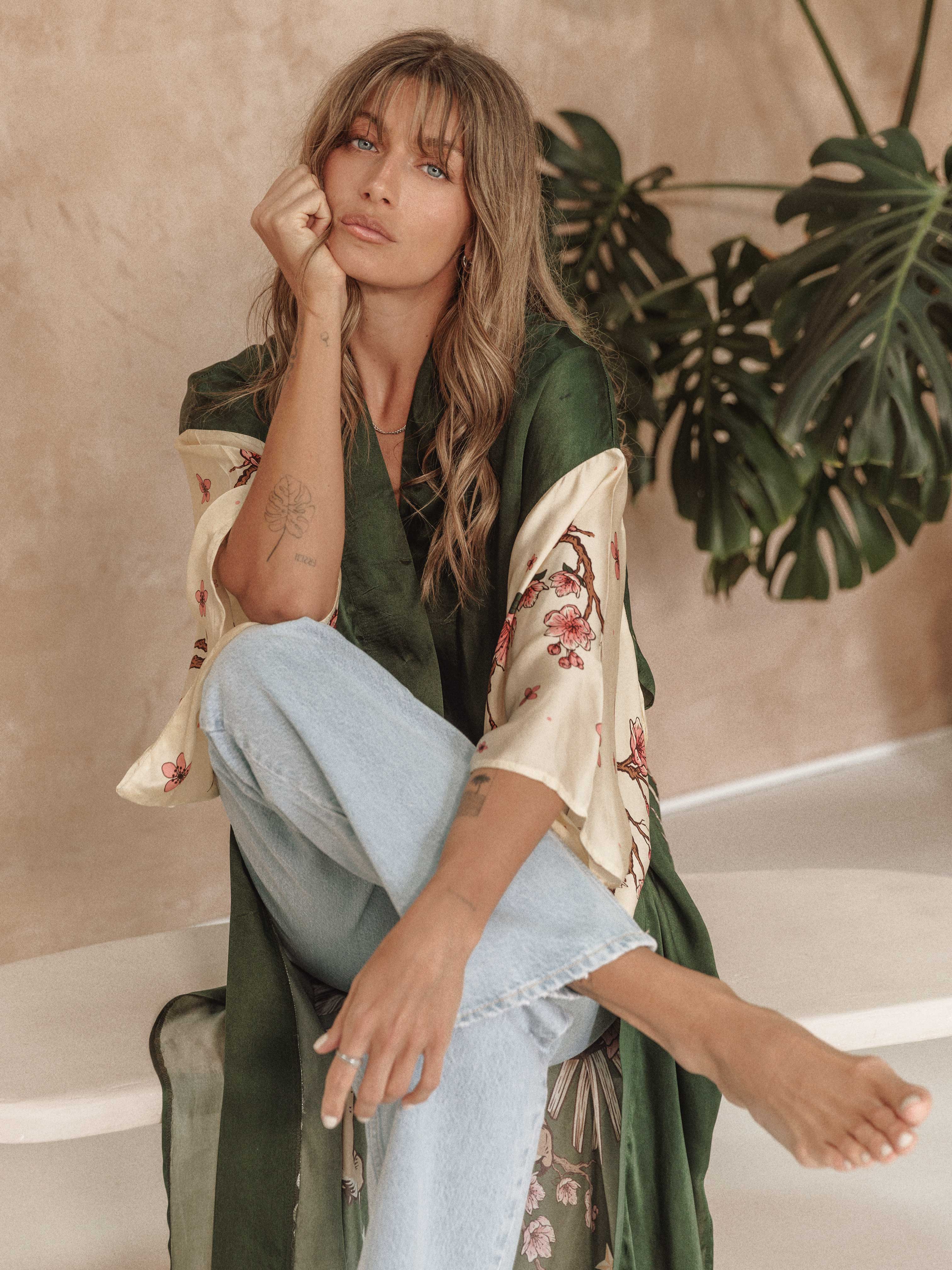 Woman wearing a dark green kimono-style cardigan with floral print sleeves, paired with light-wash jeans, seated indoors.