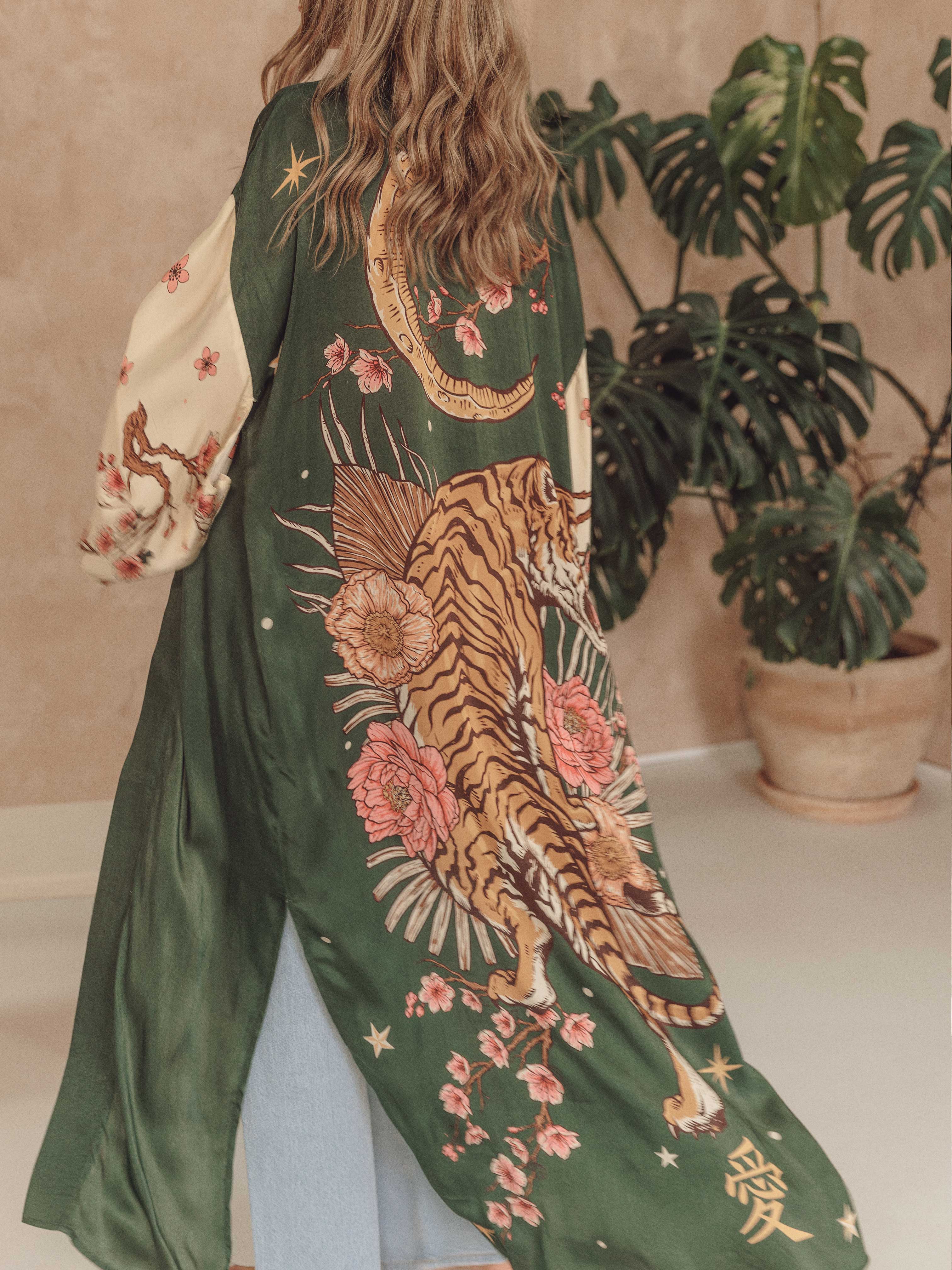 A woman viewed from behind wearing a long, flowing green kimono-style cardigan with a large tiger, moon, and floral print. The cardigan has cream-colored sleeves with pink floral patterns and is styled in an indoor setting with a potted plant in the background.
