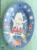 An oval ceramic plate with a hand-painted design featuring a white hand holding a silver bird, surrounded by pink protea flowers, green leaves, and white stars on a dusty blue background, with a banner reading 'LOVE'.