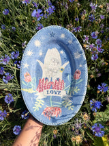 An oval blue ceramic plate with a hand-painted white bird, flowers, and the word 'LOVE', held by a hand against a background of green grass and purple wildflowers.