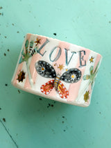 A handmade ceramic candle holder with a pink and white background, decorated with a black and red butterfly, gold stars, and the word 'LOVE' in blue letters. The candle is shown in against a turquoise blue surface