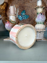 A handmade ceramic candle holder with a pink and white background, decorated with a black and red butterfly, gold stars, and the word 'LOVE' in blue letters. The candle is shown in a styled setting with other vintage bird figurines and decorative items.