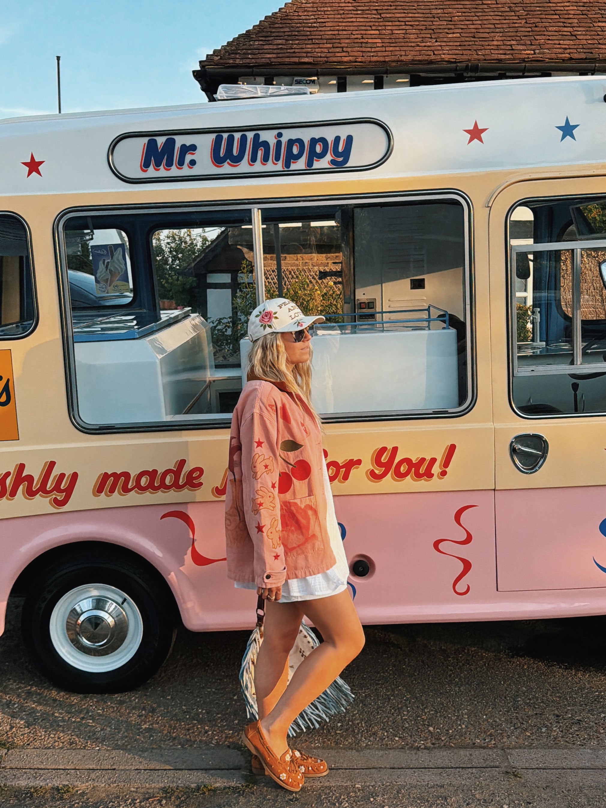 Woman wearing a pink hand-painted jacket with cherry, flower, heart, and bunny artwork while walking past a vintage Mr Whippy ice cream van.