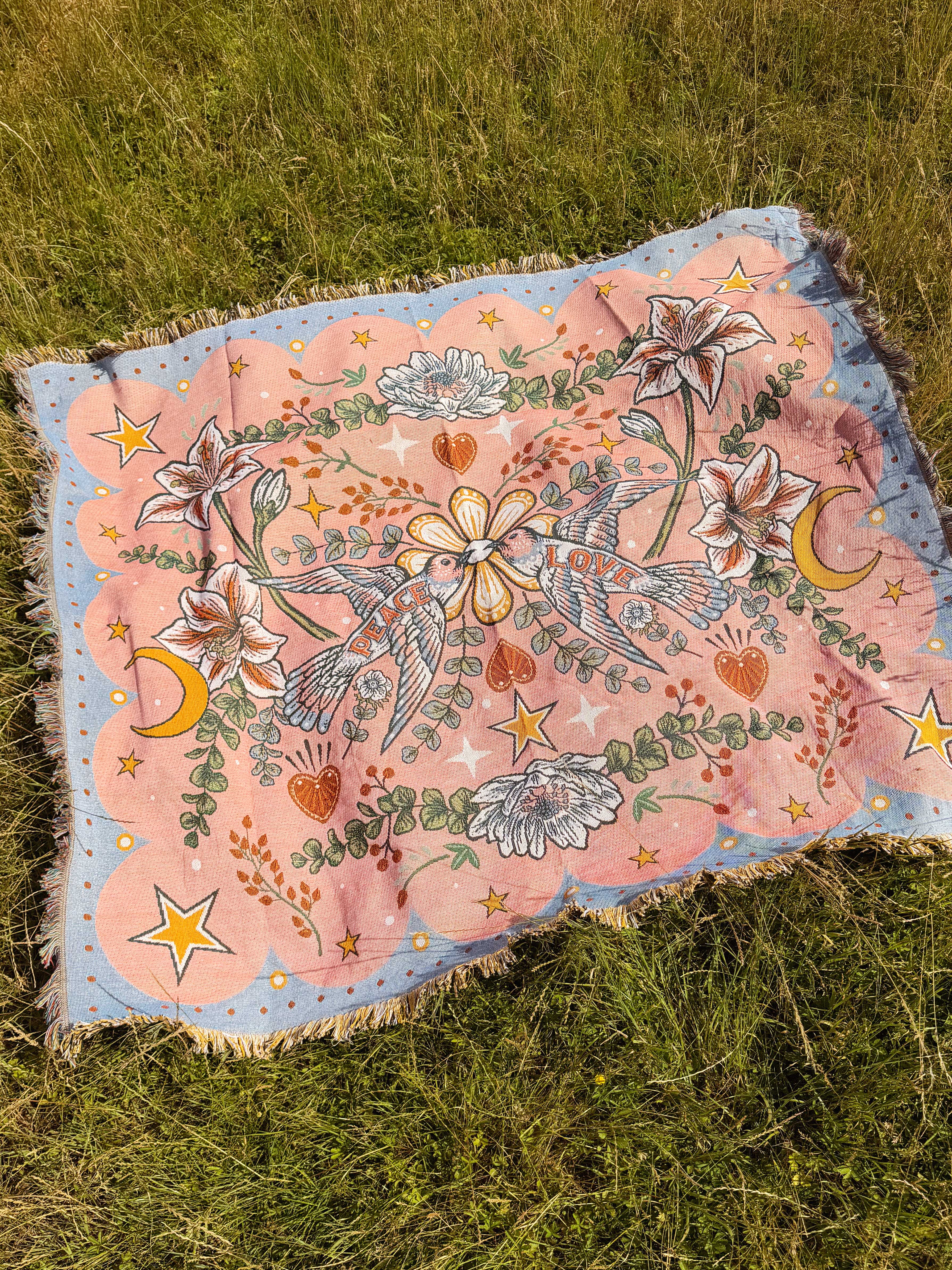 Pink woven throw blanket with hummingbirds, lilies, stars, and moons spread out on grass