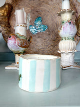 A ceramic candle holder with vertical light blue and white stripes in the foreground, flanked by two vintage-style bird candlesticks against a rustic woven backdrop.