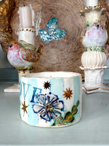 A handmade ceramic candle holder with a light blue and white striped body, decorated with a blue flower, green leaves, gold stars, and the letters 'WE' in blue, displayed on a shelf between other decorative bird and birdhouse figurines.