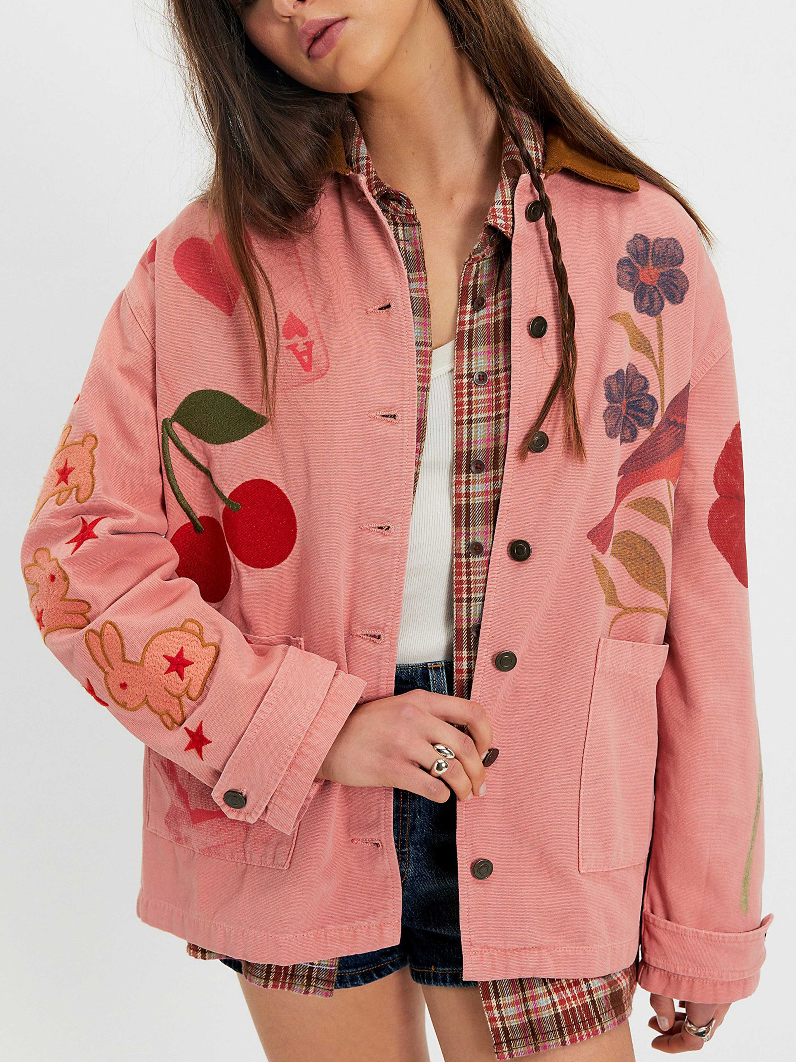 Woman wearing a pink hand-painted jacket featuring cherry, flower, heart, and bunny artwork over a plaid shirt.