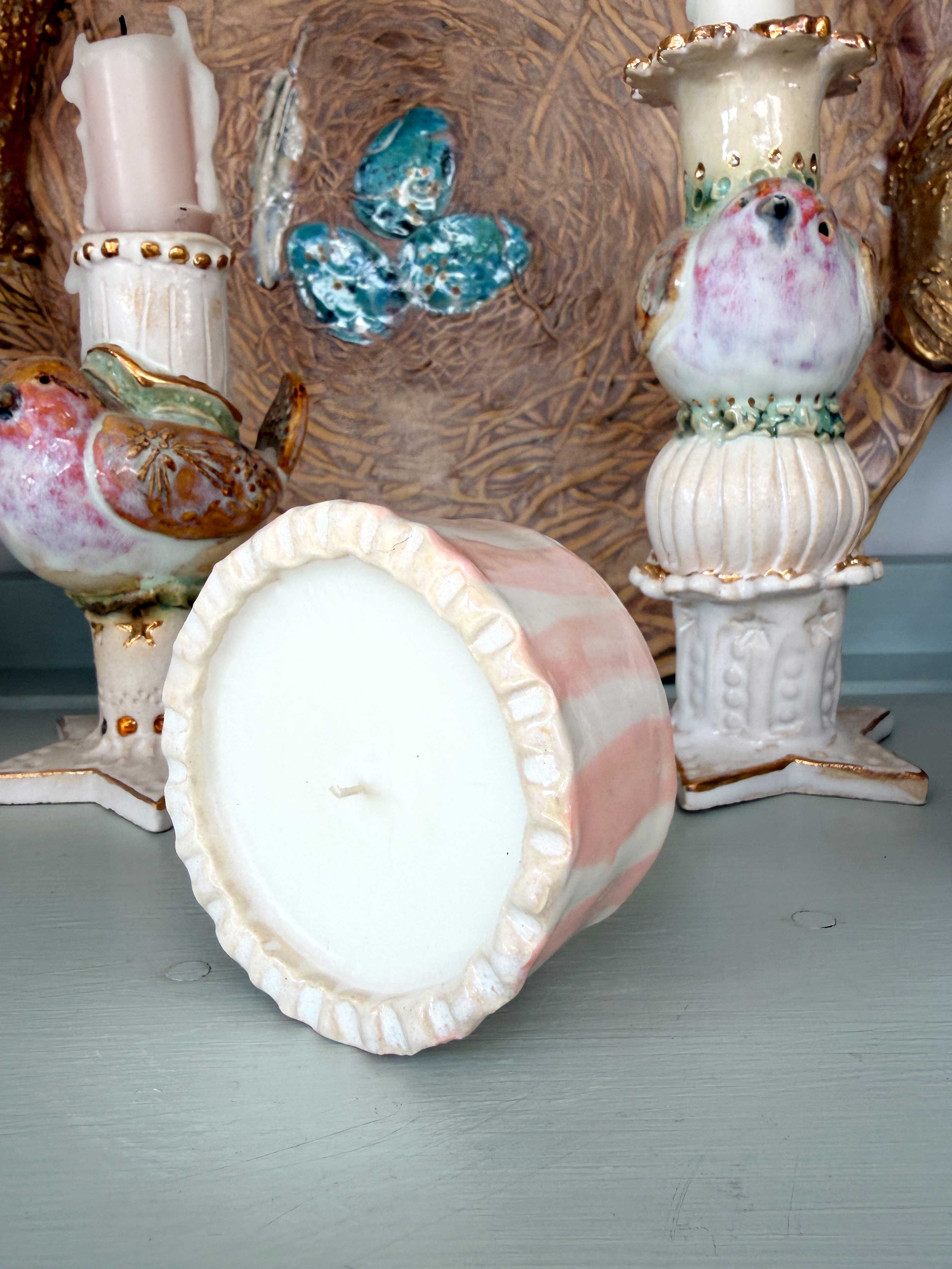 A pink and white marbled ceramic candle holder with a ruffled rim, shown tipped over on its side to display the white wax pool inside. The background features other decorative ceramic bird and butterfly figurines.