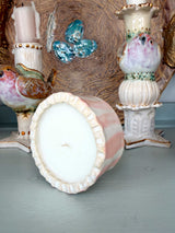 A pink and white marbled ceramic candle holder with a ruffled rim, shown tipped over on its side to display the white wax pool inside. The background features other decorative ceramic bird and butterfly figurines.