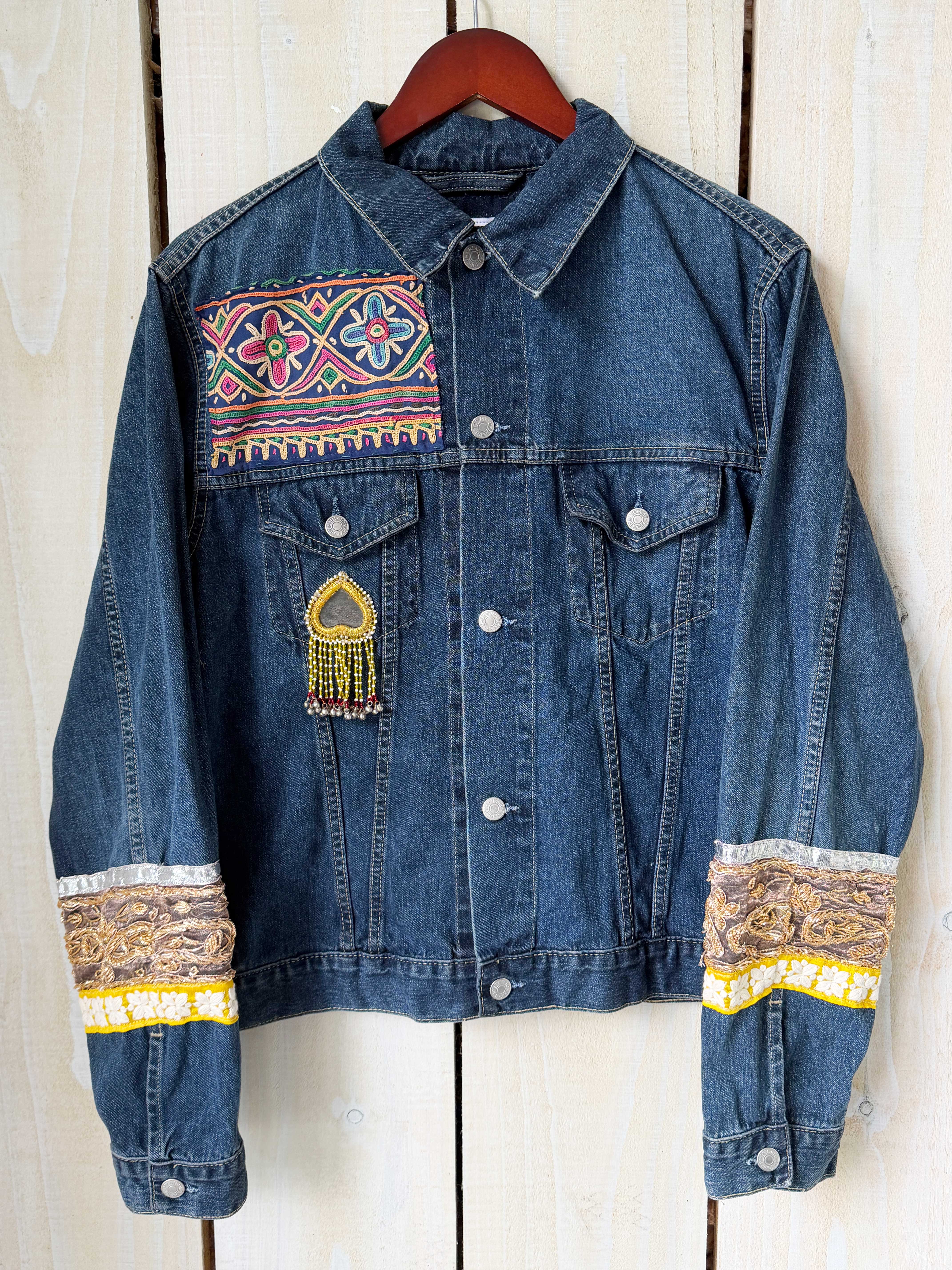 Front view of a blue denim jacket with colourful embroidered arms hanging on a white wooden rustic wall