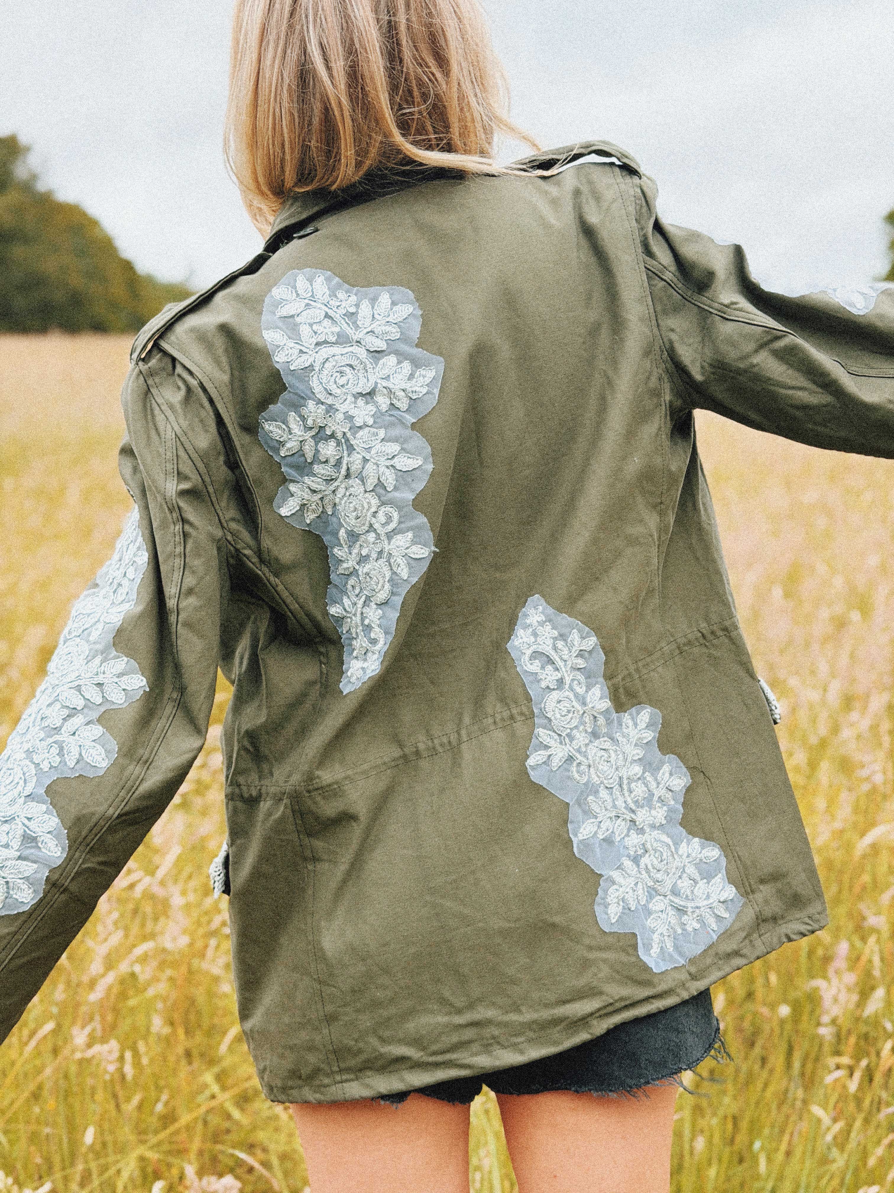 Olive green military jacket with white floral lace appliqué and decorative trim, back view.