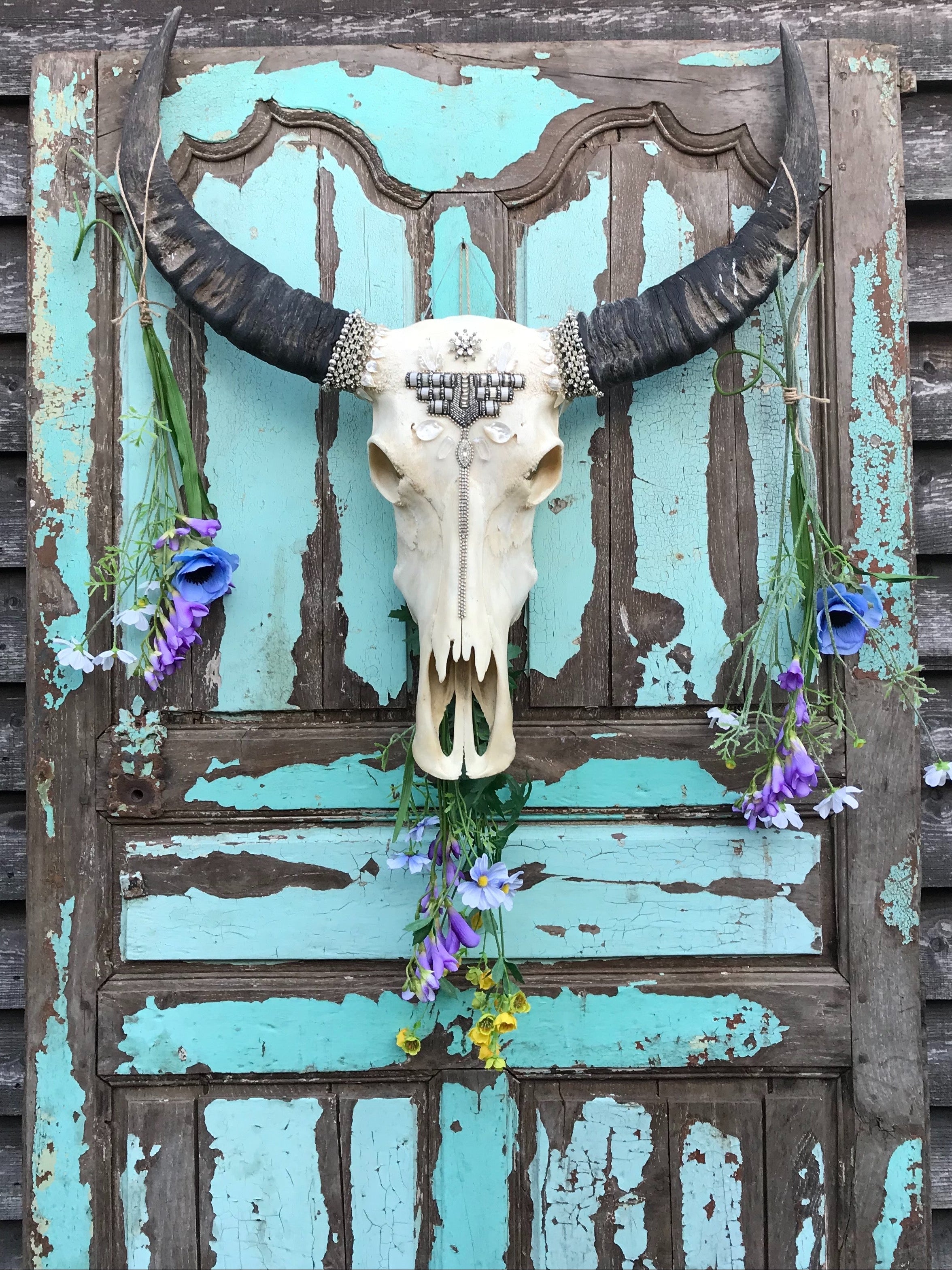 Decorative Thai buffalo skull wall art with natural black horns, silver crystal beadwork, shell embellishments, and cascading wildflowers displayed on a distressed turquoise vintage door.