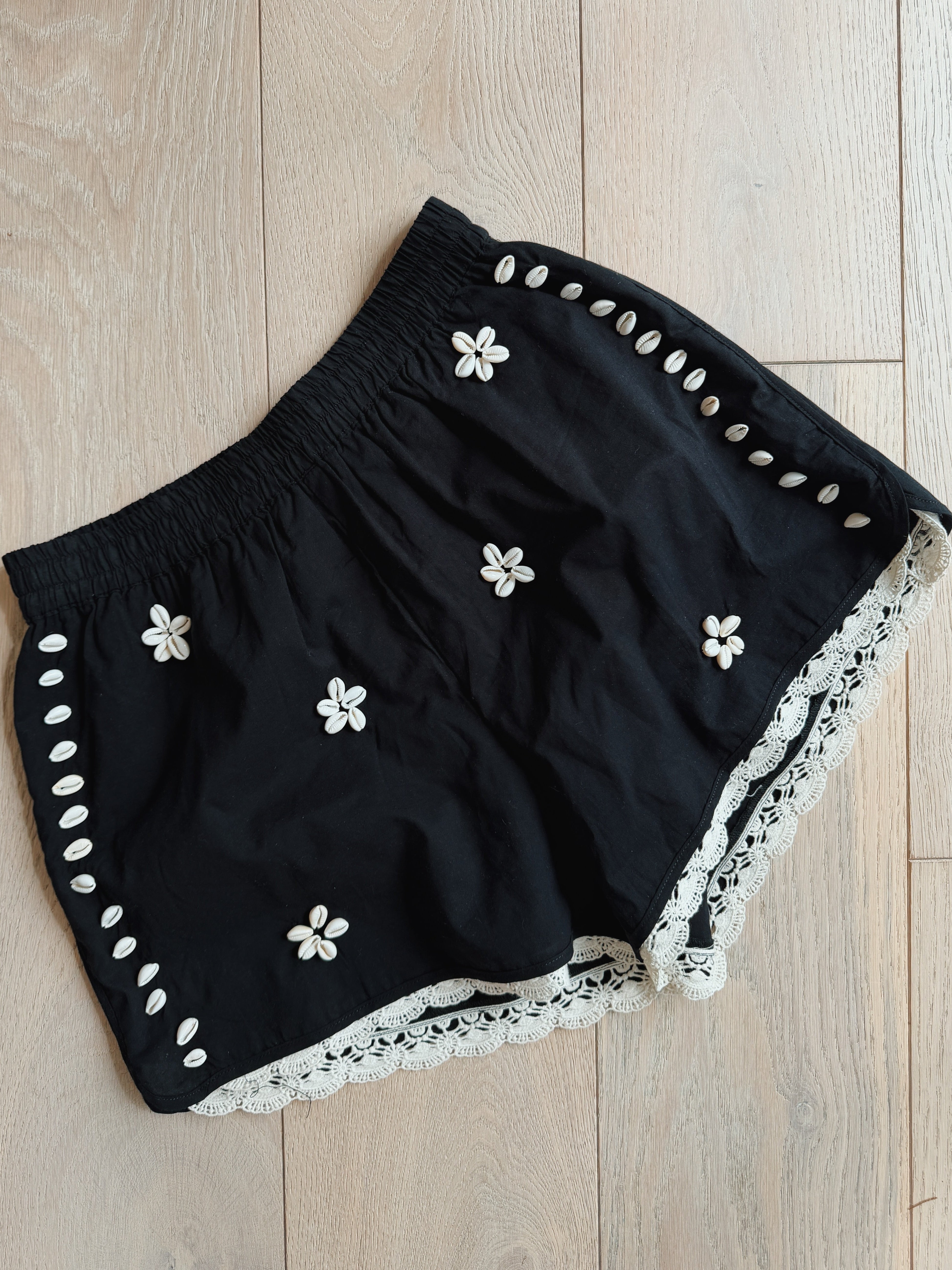 Black shorts with white floral embroidery, shell side trim, scalloped lace hem, and an elastic waistband, displayed flat on a light wood floor.