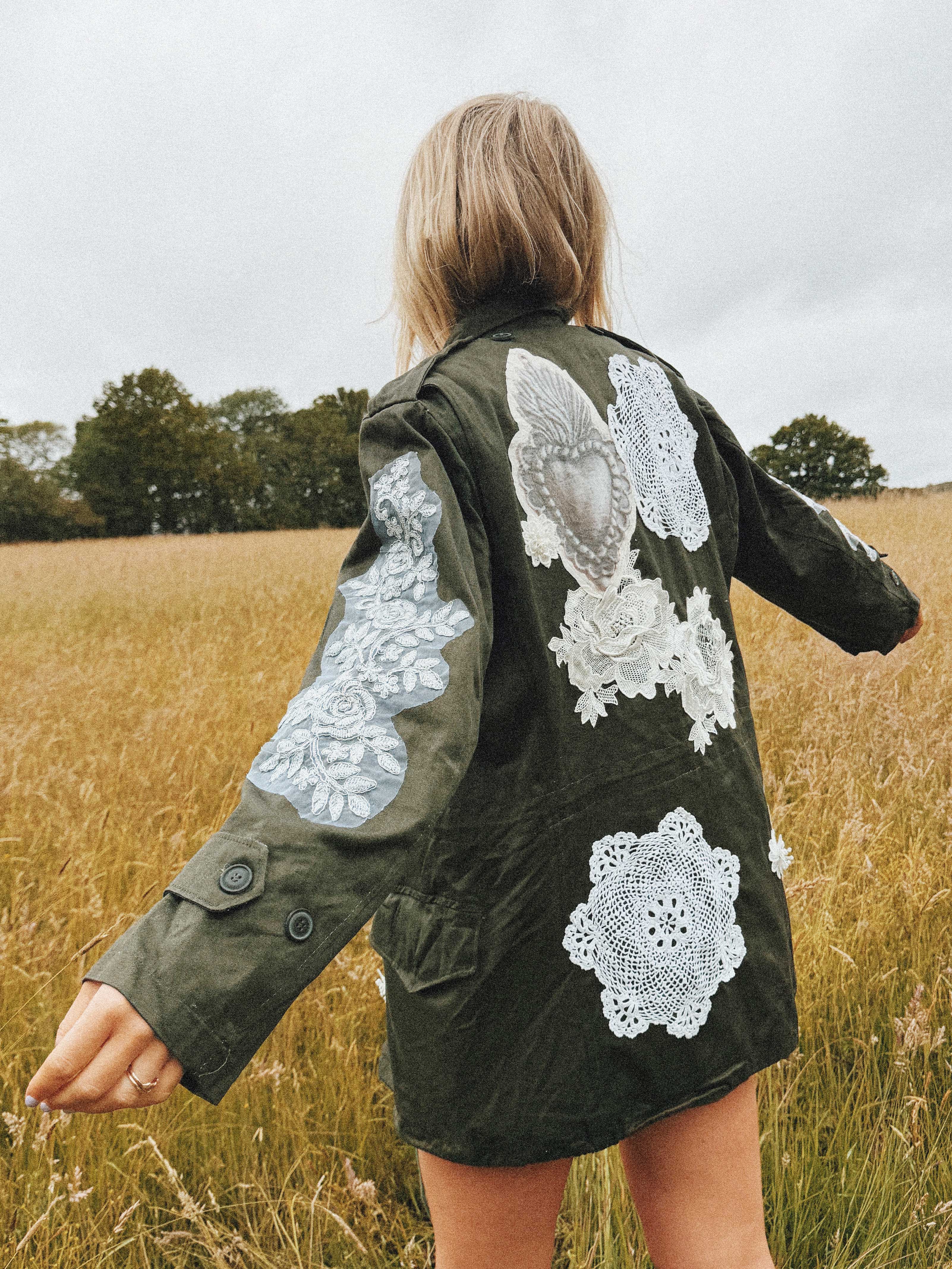 Olive green military jacket with white floral lace appliqué on the back and sleeves, back view.