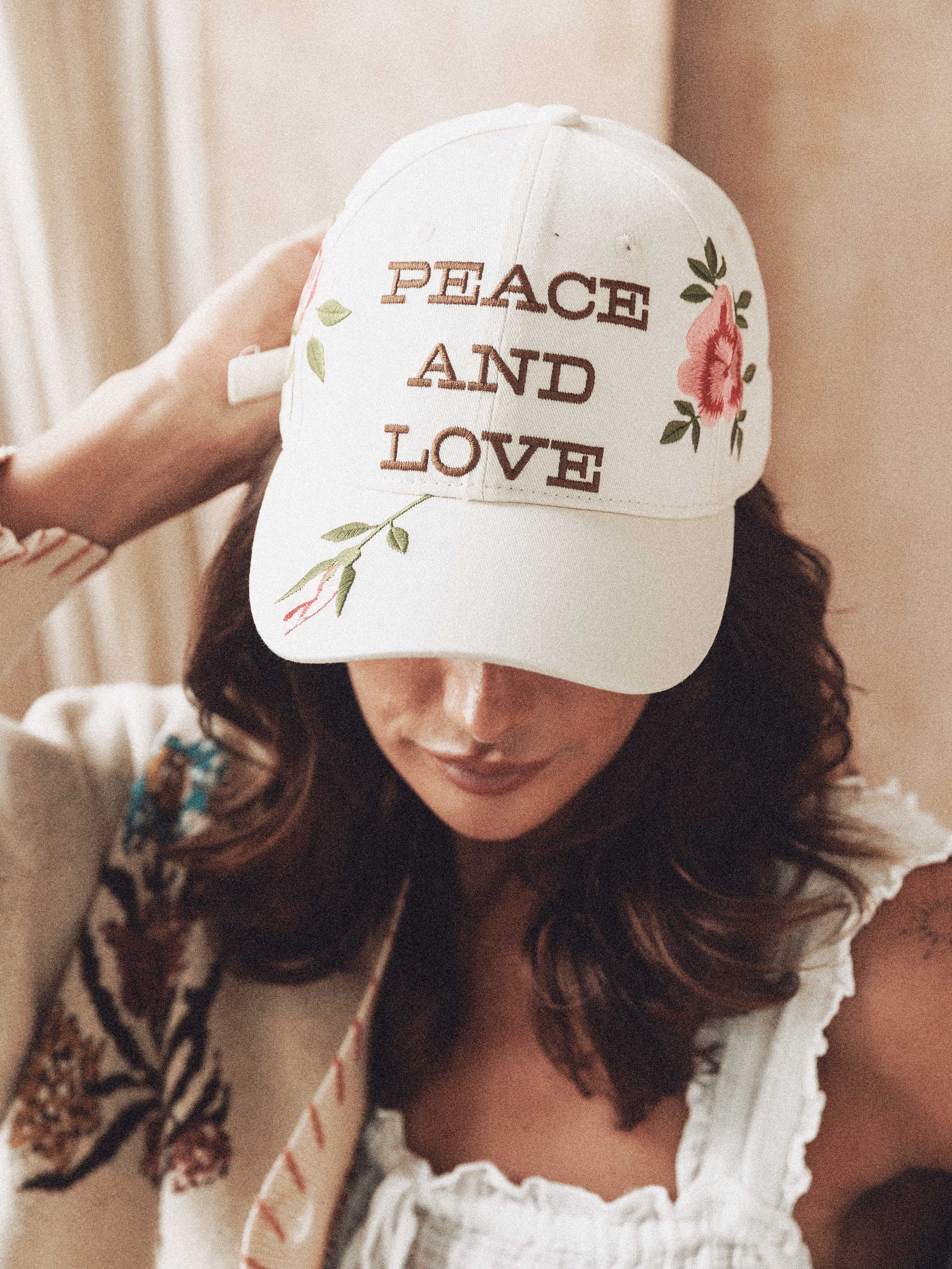 Woman wearing a cream baseball cap embroidered with "Peace and Love" and pink roses.
