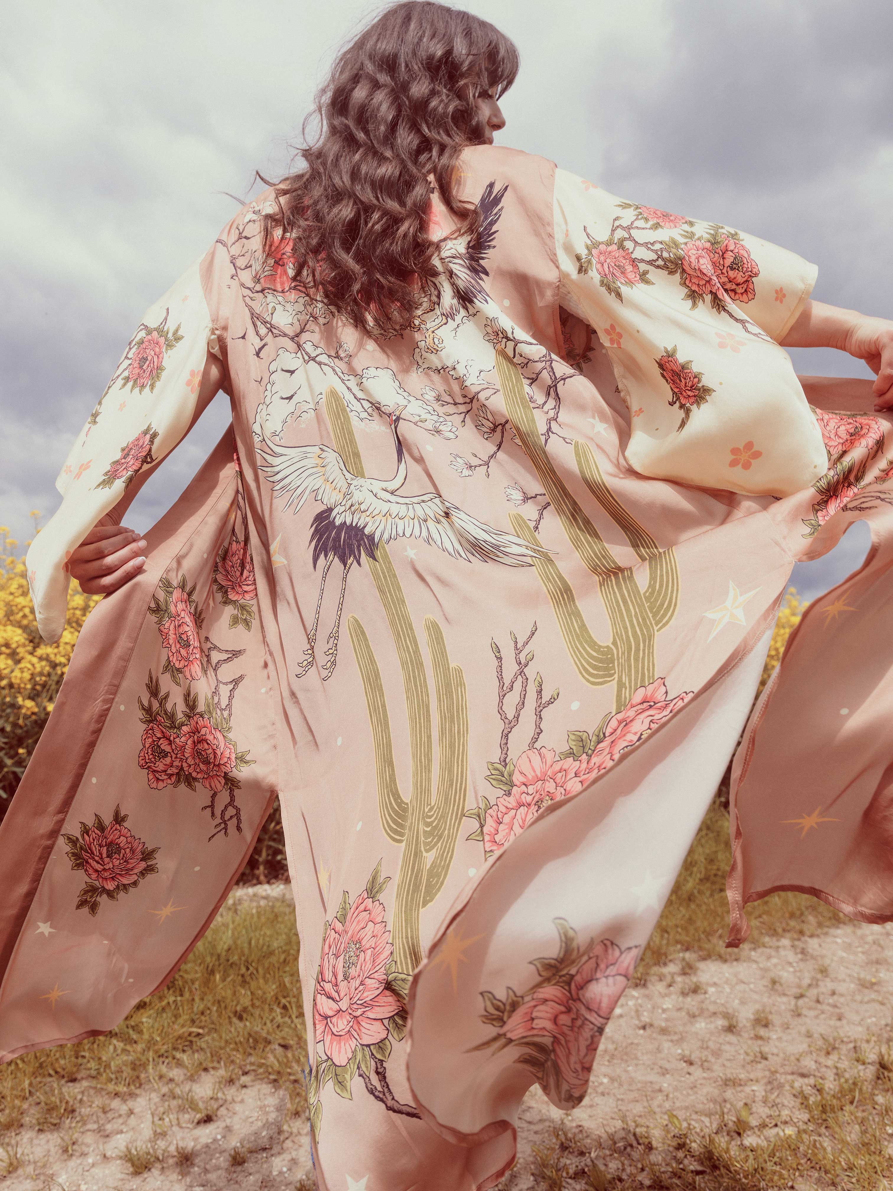 Person wearing a blush pink kimono-style robe featuring desert cacti, cranes, blooming flowers, and flowering branches while standing outdoors.