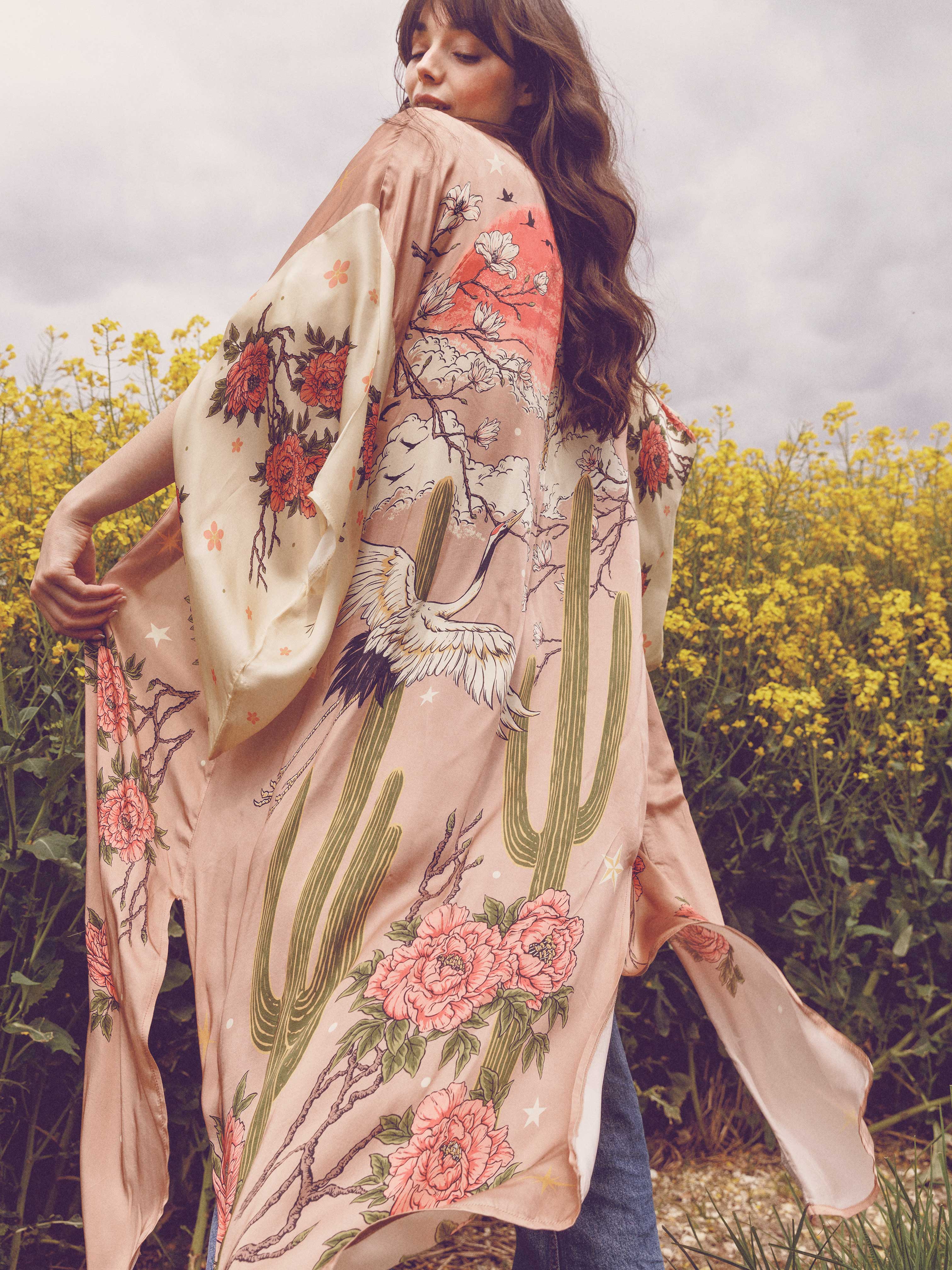 Person wearing a blush pink kimono-style robe featuring desert cacti, cranes, blooming flowers, and flowering branches while standing outdoors.