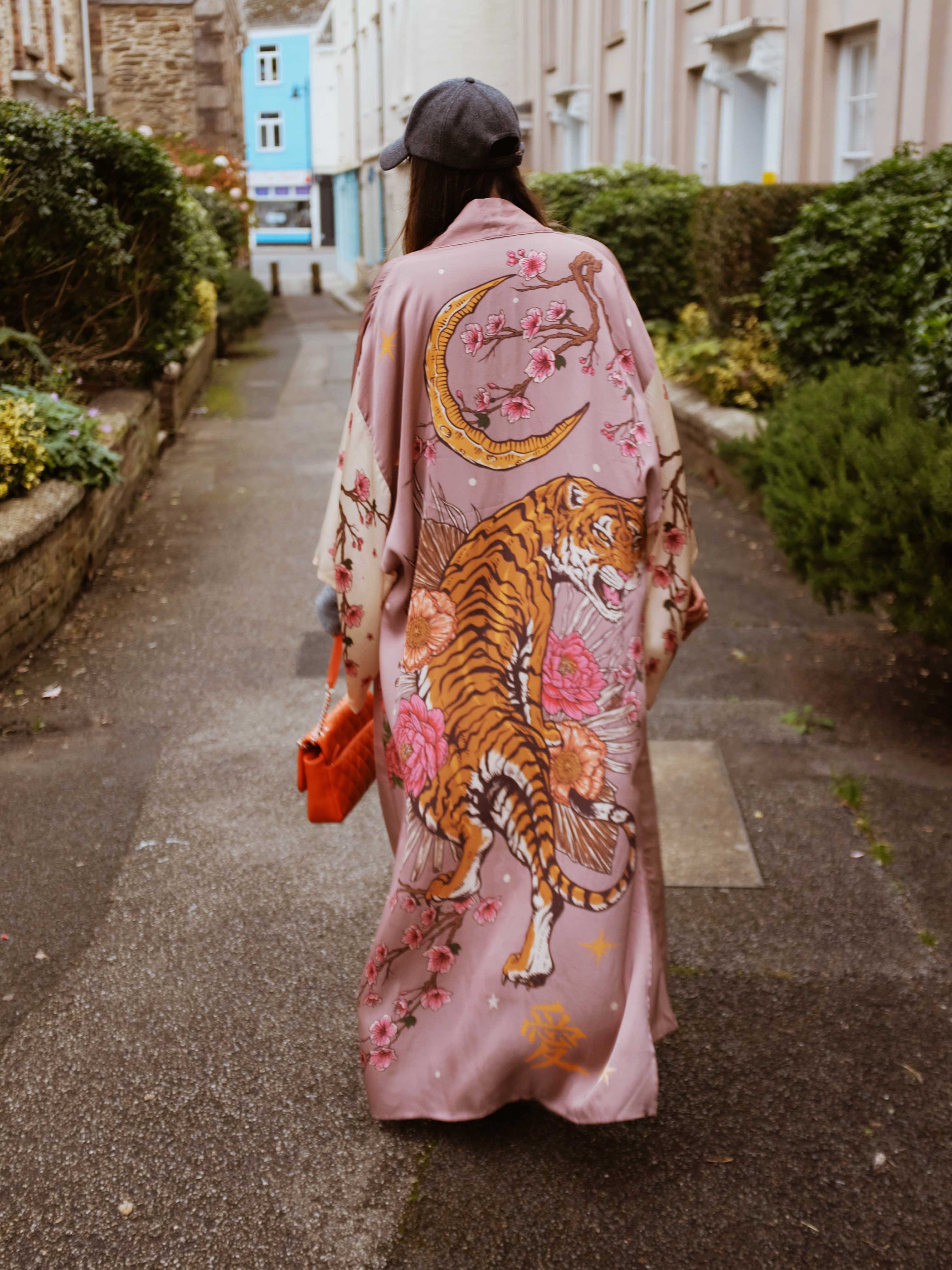 Person wearing a long pink kimono-style robe featuring a tiger, crescent moon, cherry blossoms, and floral print on the back while walking outdoors.