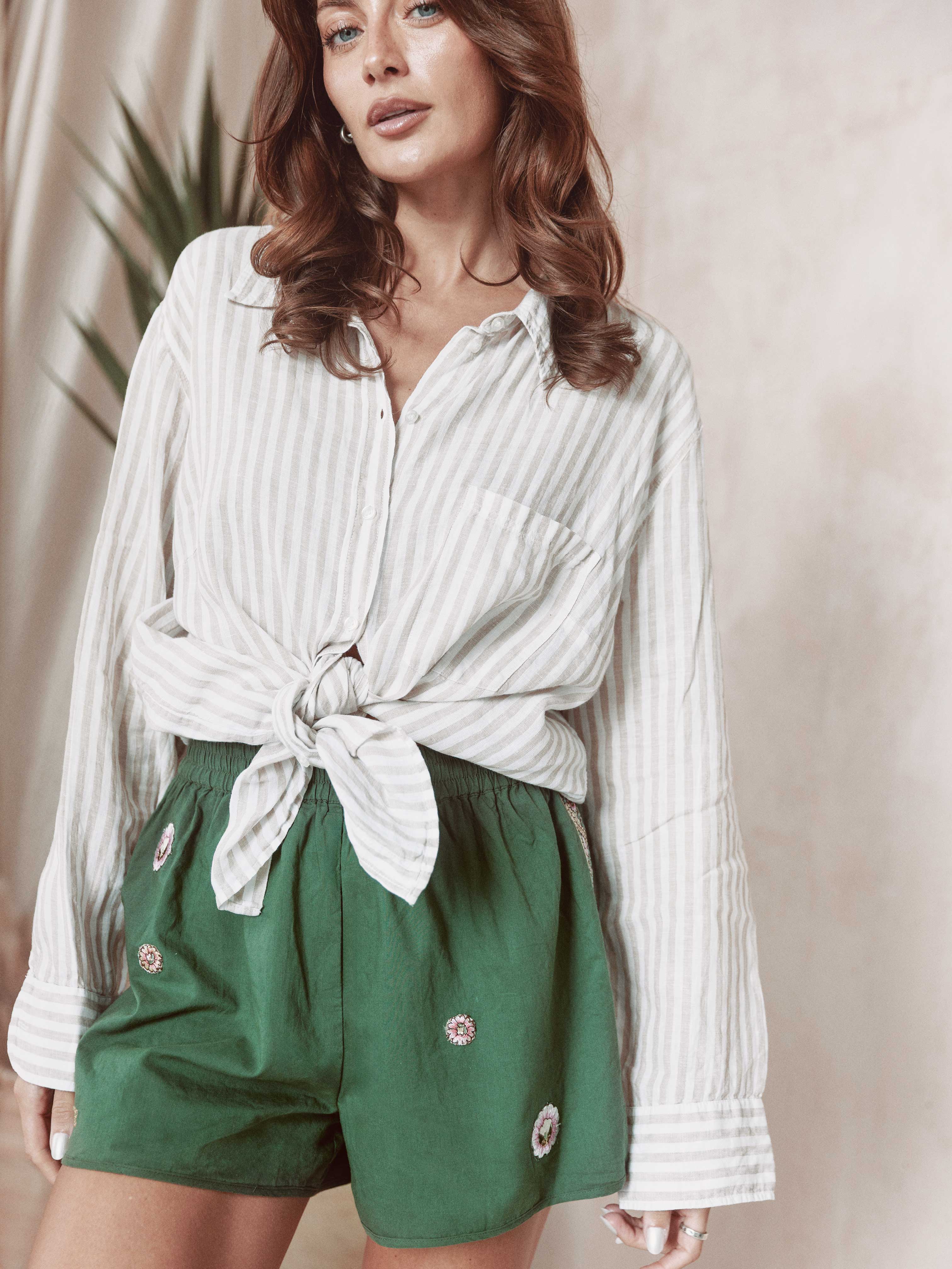 Person wearing green shorts with floral embroidered details and decorative side trim, paired with a white striped button-up shirt.