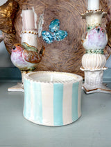 A handmade ceramic candle holder with vertical light blue and white stripes, featuring a scalloped rim and a single white candle inside, displayed on a light blue-gray surface with decorative bird and birdhouse figurines in the background.