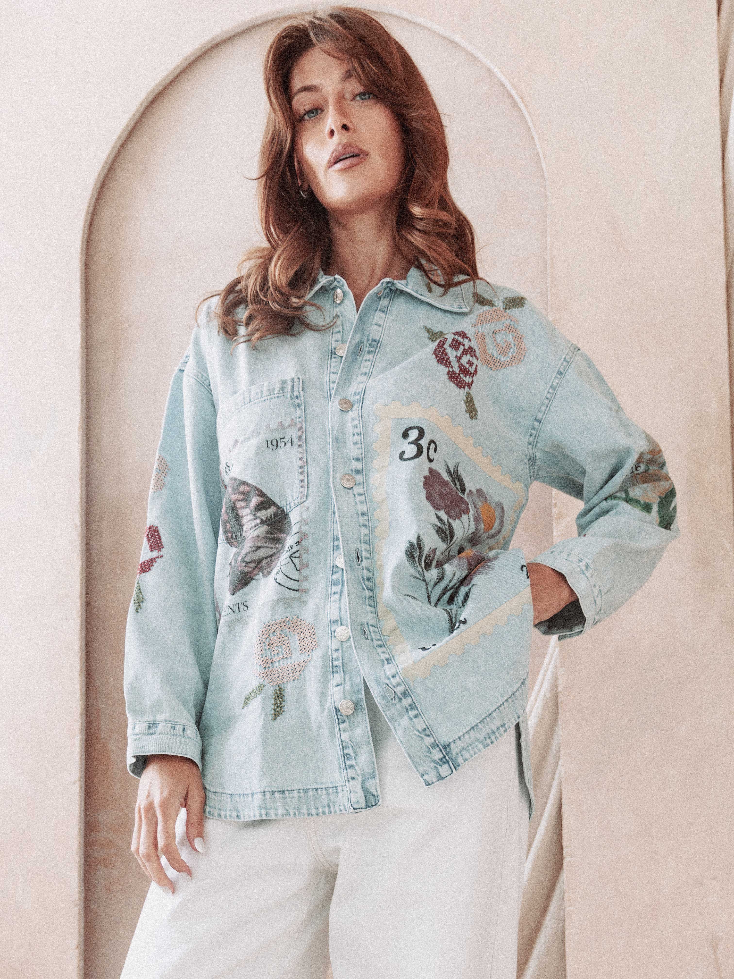 Front view of a person wearing a light wash denim button-up shirt featuring vintage-inspired postage stamp patchwork and floral cross-stitch embroidery, paired with white pants.