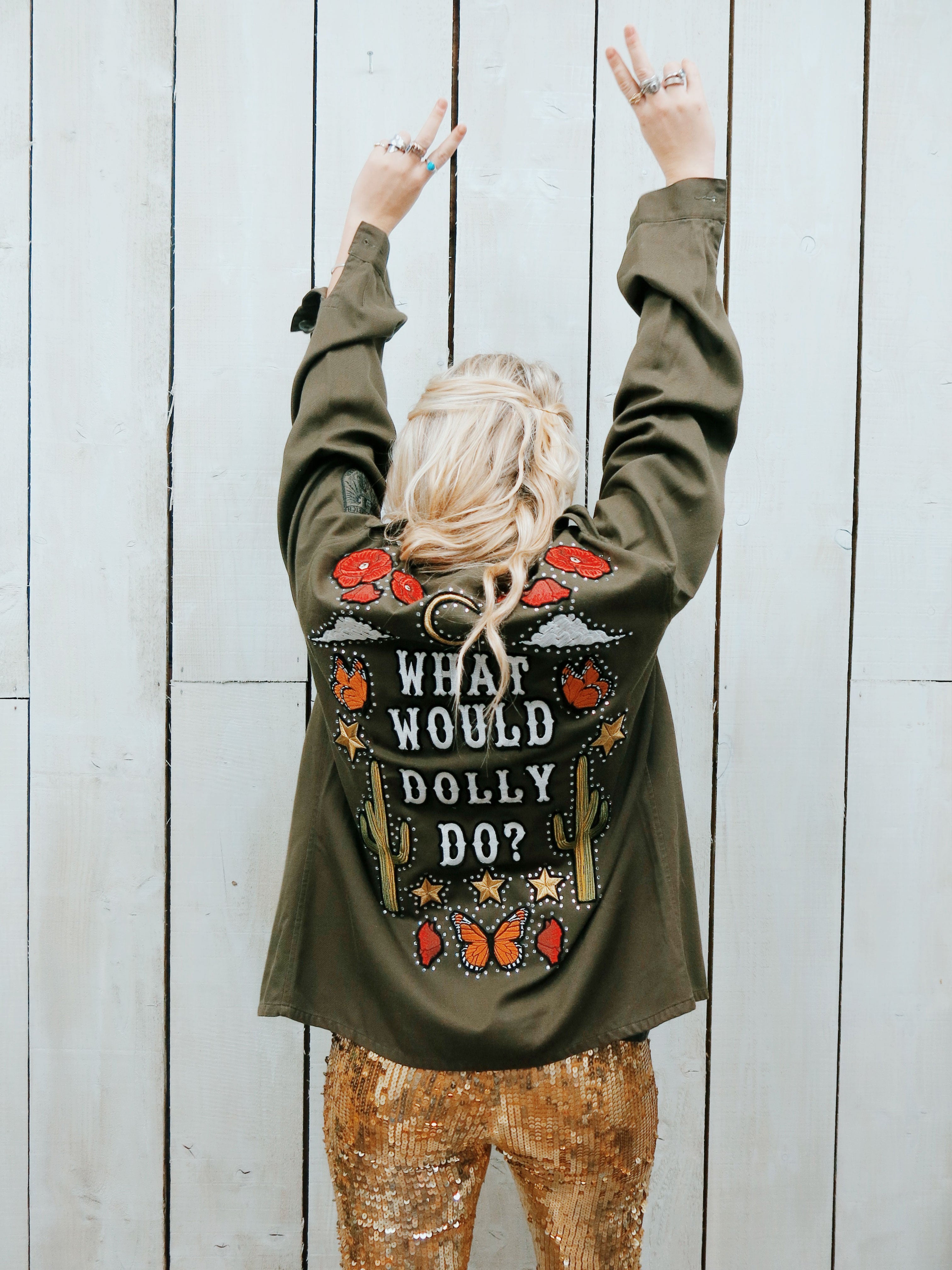 Back view of a woman styling the What Would Dolly Do embroidered khaki army jacket with gold sequin trousers, showing its western motifs and rhinestones.