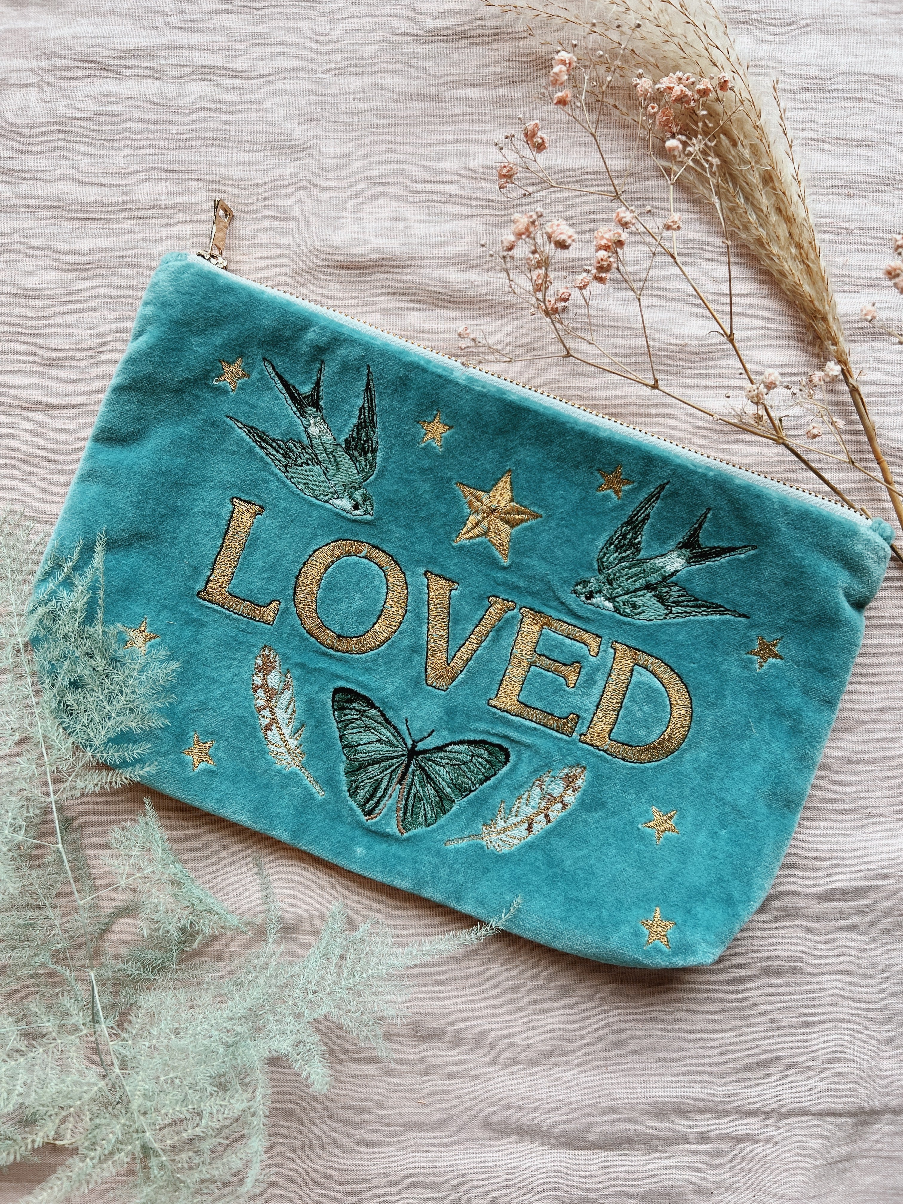 Turquoise velvet zip pouch embroidered with the word "LOVED," swallows, butterflies, feathers, and stars, styled with dried flowers.