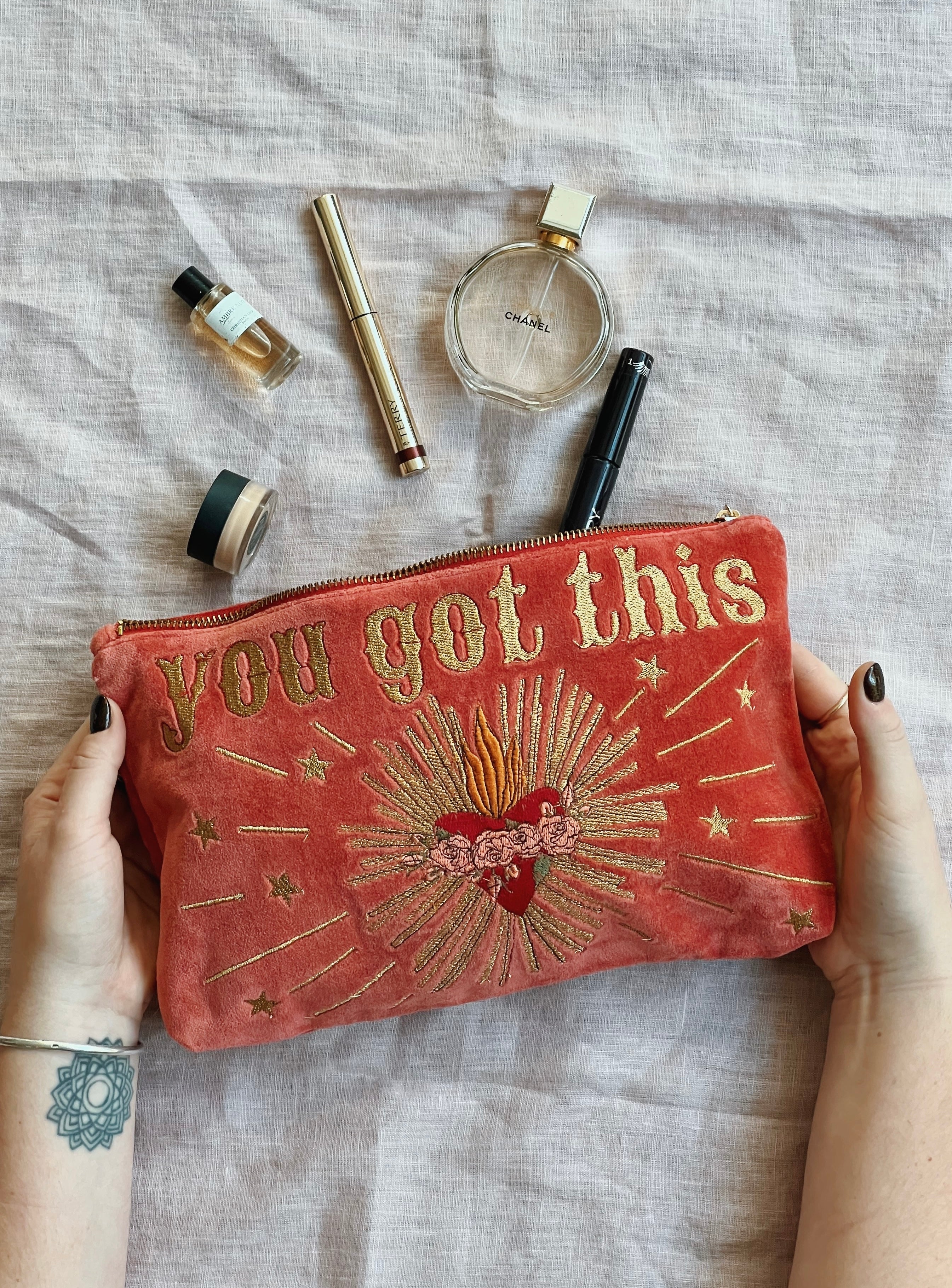 Hands holding a rich pink velvet zip pouch embroidered with the phrase "you got this" and a radiant sacred heart design, surrounded by everyday beauty essentials.