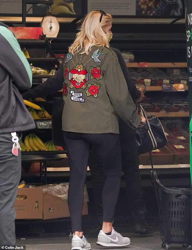 A woman shopping in a grocery store wearing an olive green military jacket with an embroidered back design featuring a heart with clasped hands, swallows, roses, and a "TRUE LOVE" banner.