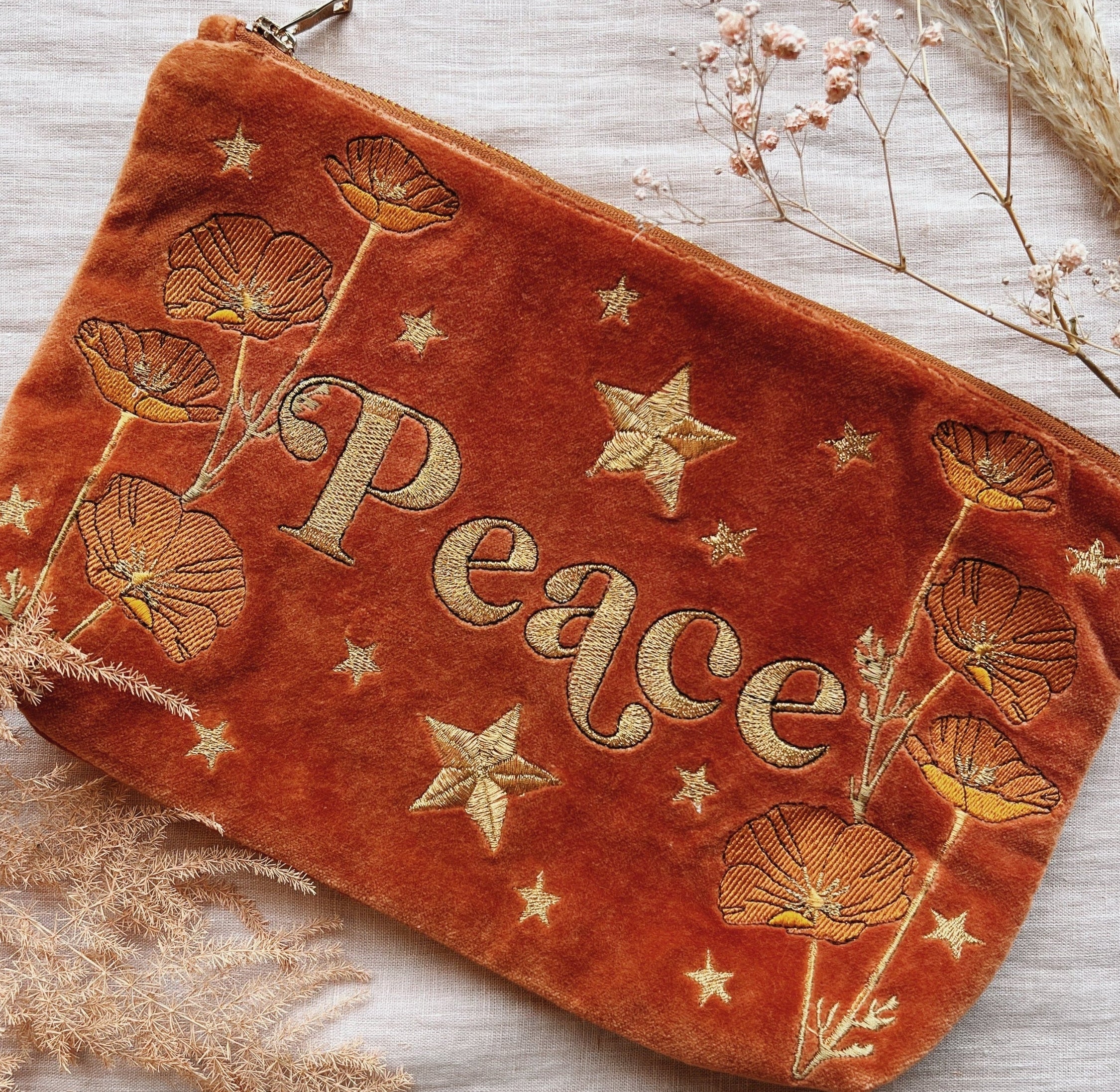 Rich rust velvet zip pouch embroidered with the word "Peace," wildflowers, and stars, styled with dried flowers.