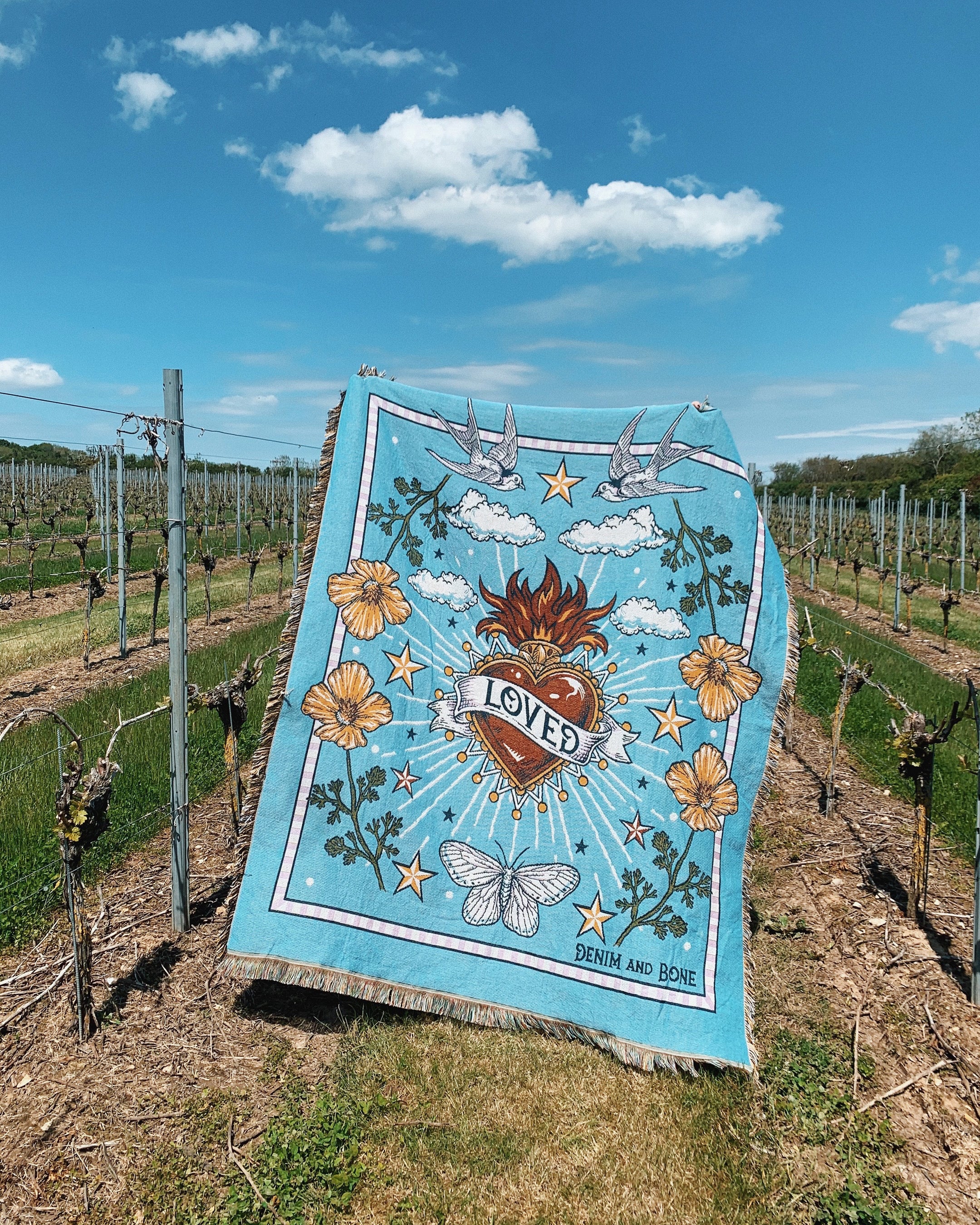 "Loved" woven throw blanket displayed outdoors in a vineyard beneath a blue sky.