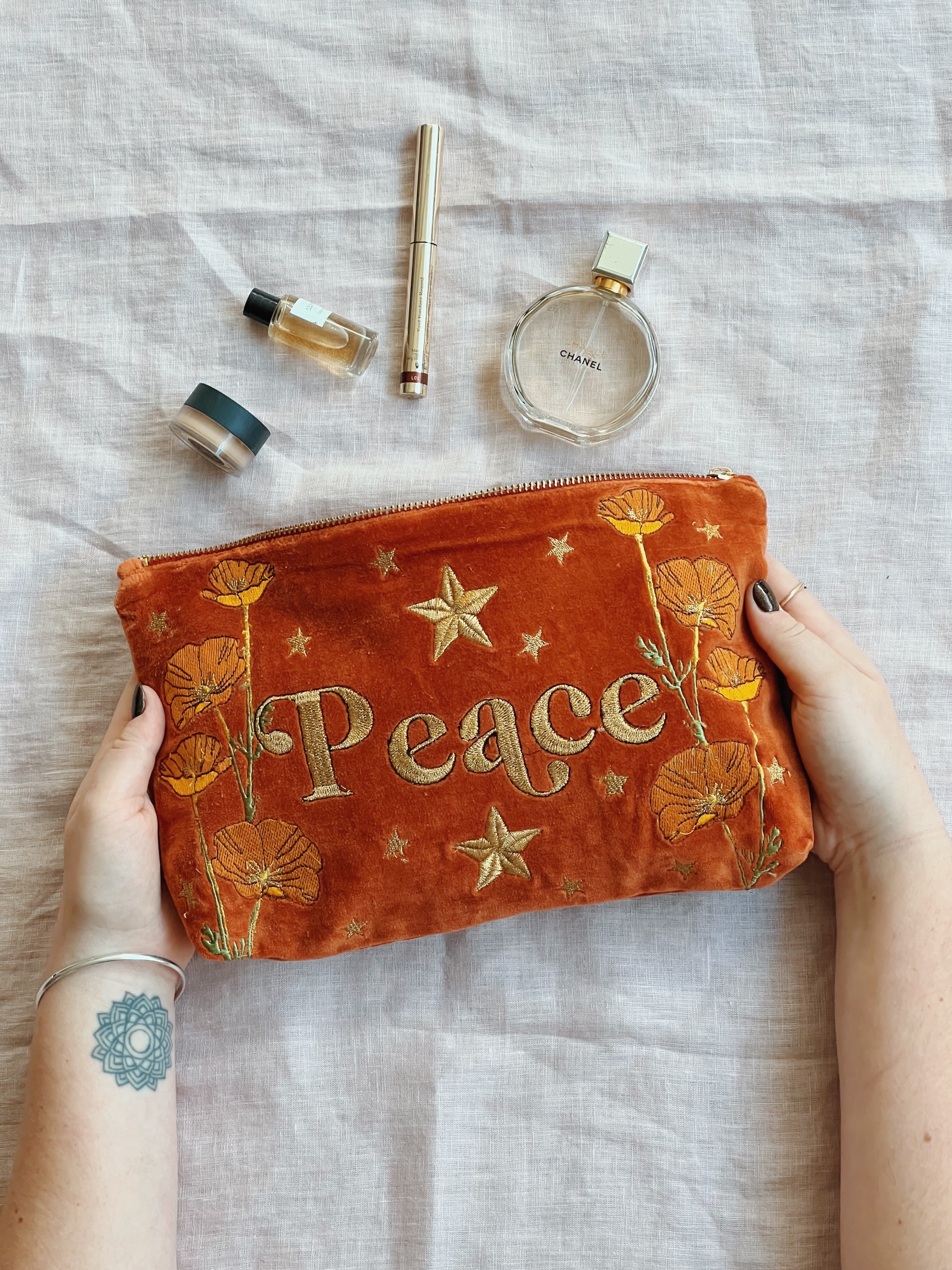 Hands holding a rich rust velvet zip pouch embroidered with the word "Peace," wildflowers, and stars, styled with beauty essentials.