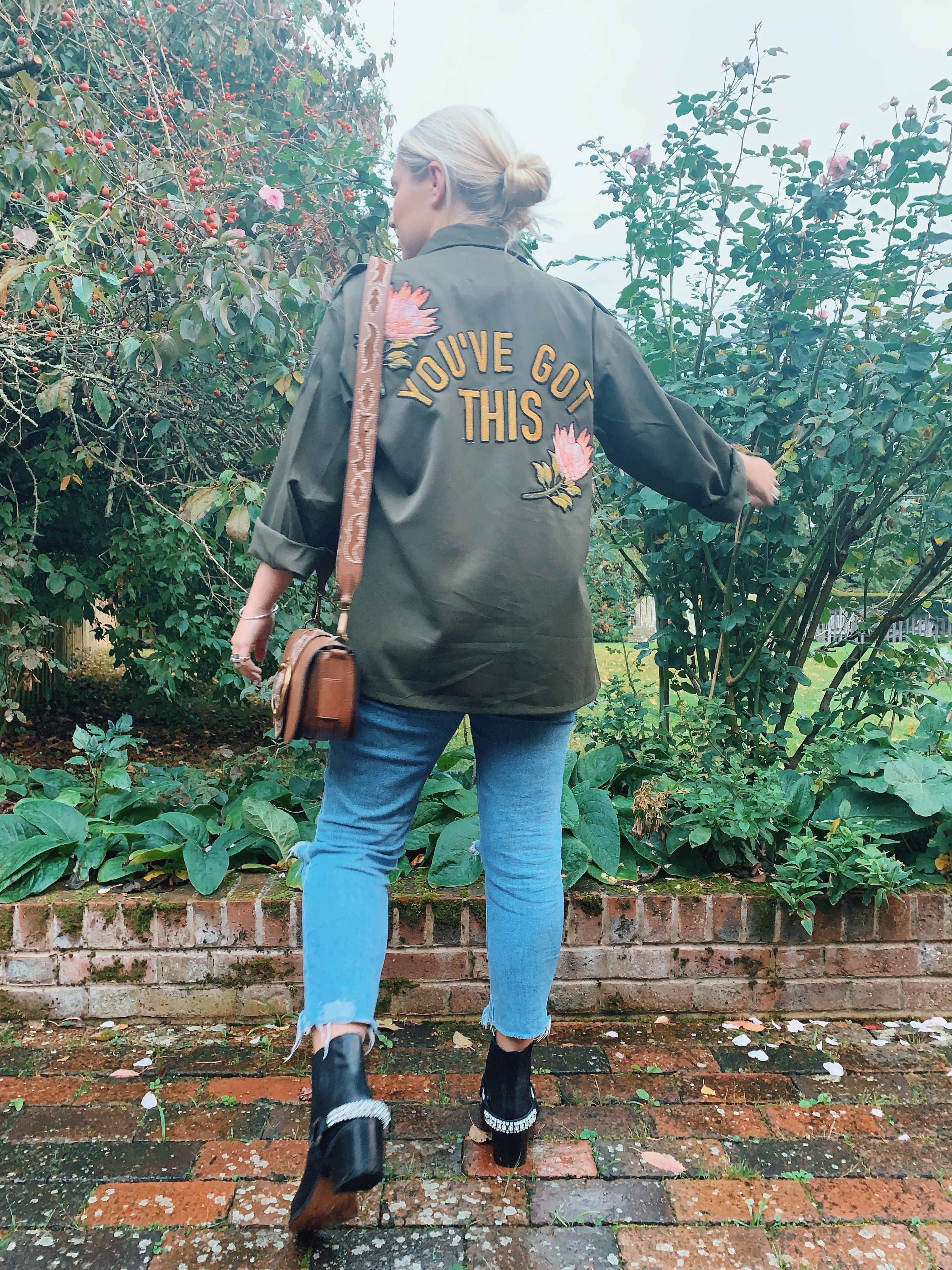 Back view of a person wearing an olive green military jacket with "YOU'VE GOT THIS" embroidery and pink protea flowers outside