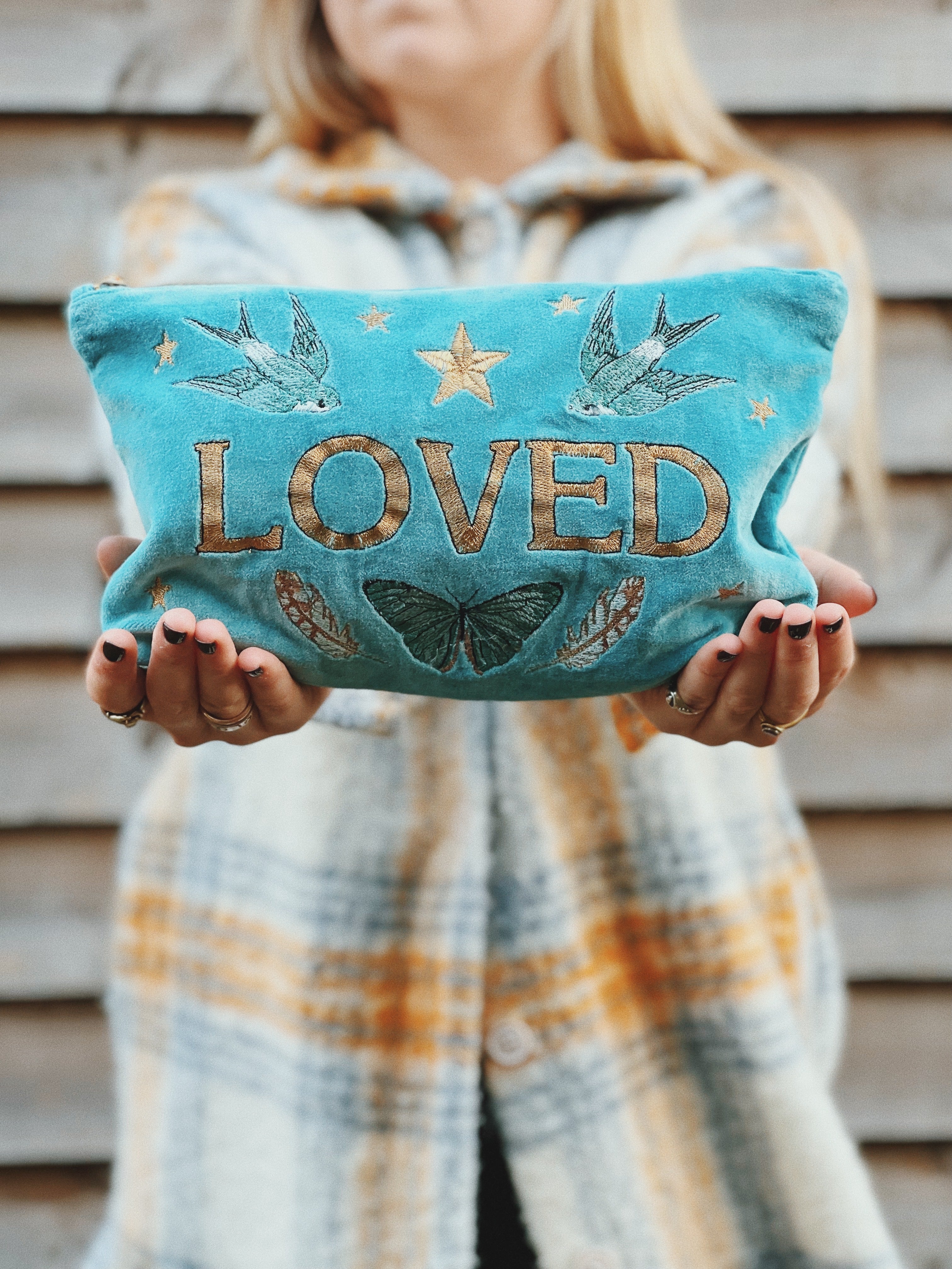 Person holding a turquoise velvet zip pouch embroidered with the word "LOVED," swallows, butterflies, feathers, and stars.