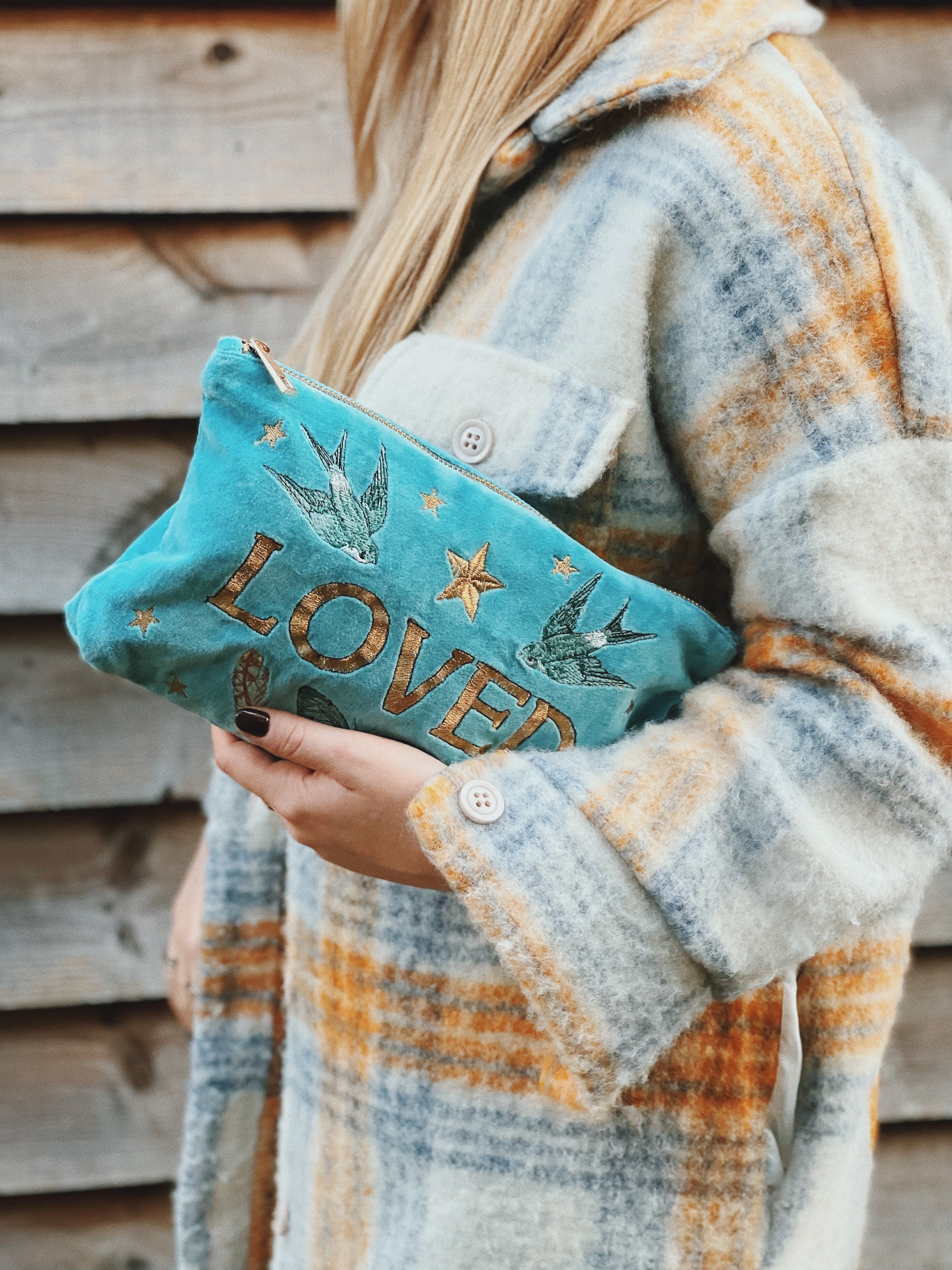 Person holding a turquoise velvet zip pouch embroidered with the word "LOVED," swallows, butterflies, feathers, and stars.