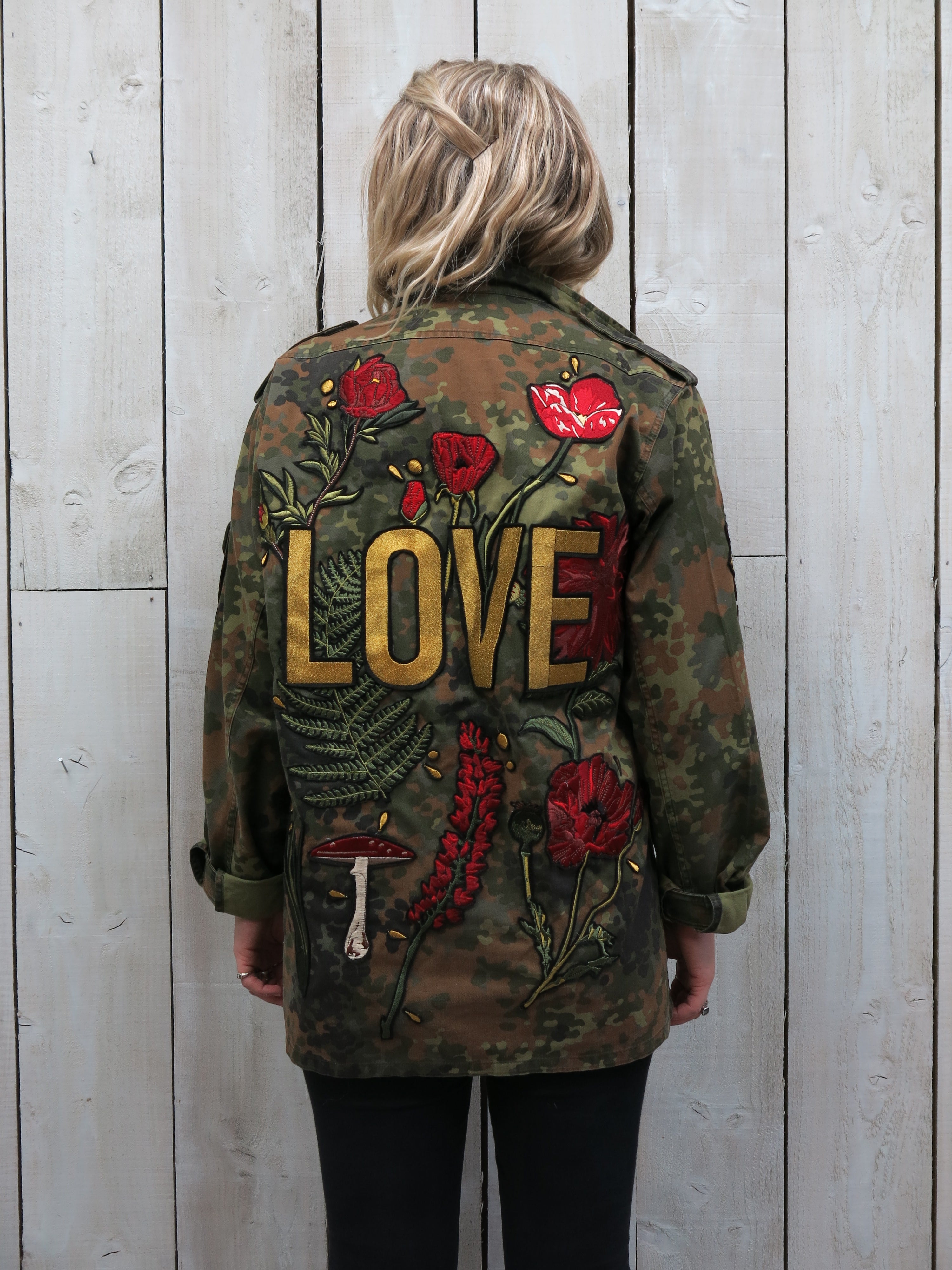 A woman viewed from behind wearing a camouflage military jacket with a large embroidered "LOVE" design surrounded by red flowers and botanical motifs.