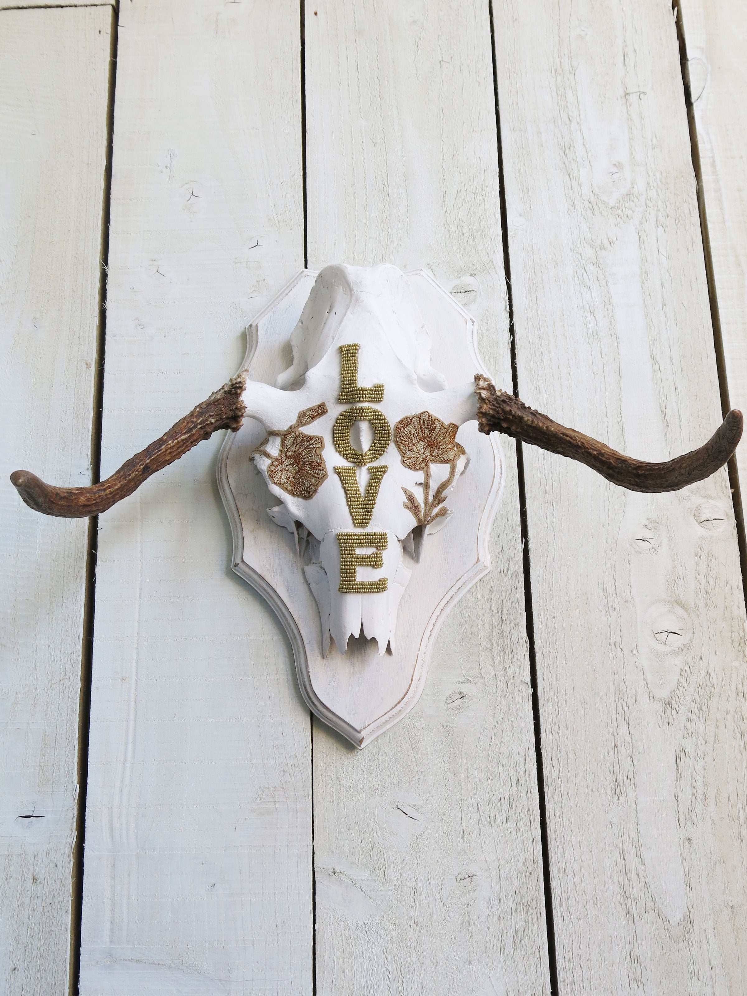 Decorative moose skull wall art with gold beaded "LOVE" lettering, floral embellishments, and natural antlers mounted on a rustic plaque.