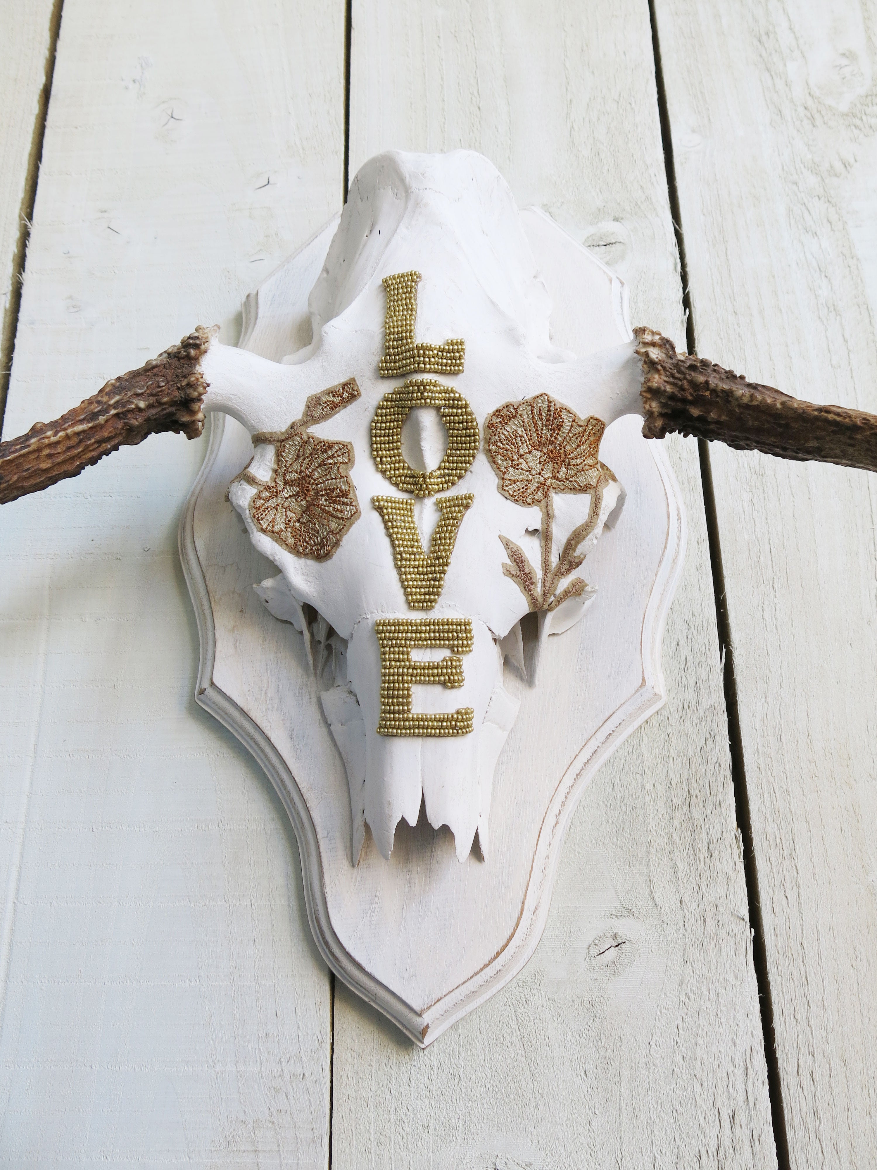 Western-inspired moose skull wall hanging featuring gold beadwork spelling "LOVE" with floral detailing.