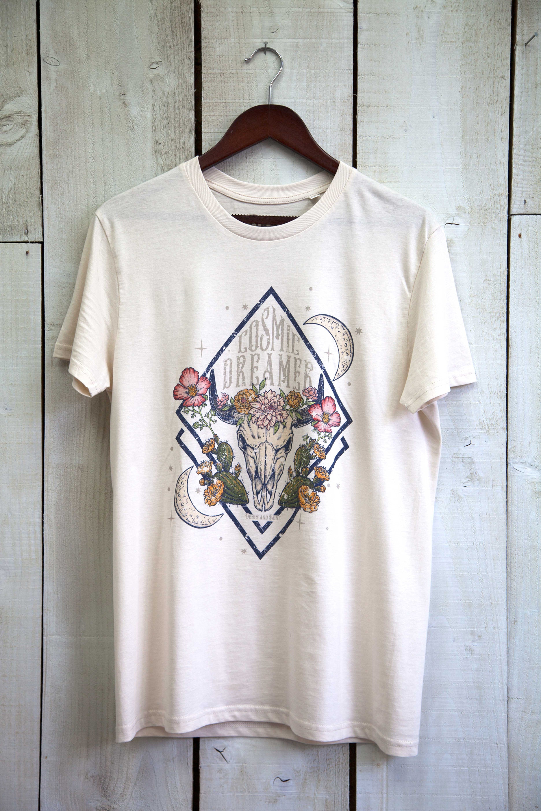 White graphic T-shirt featuring a floral longhorn skull design with crescent moons, desert cacti, and "Cosmic Dreamer" text on a rustic wood background.
