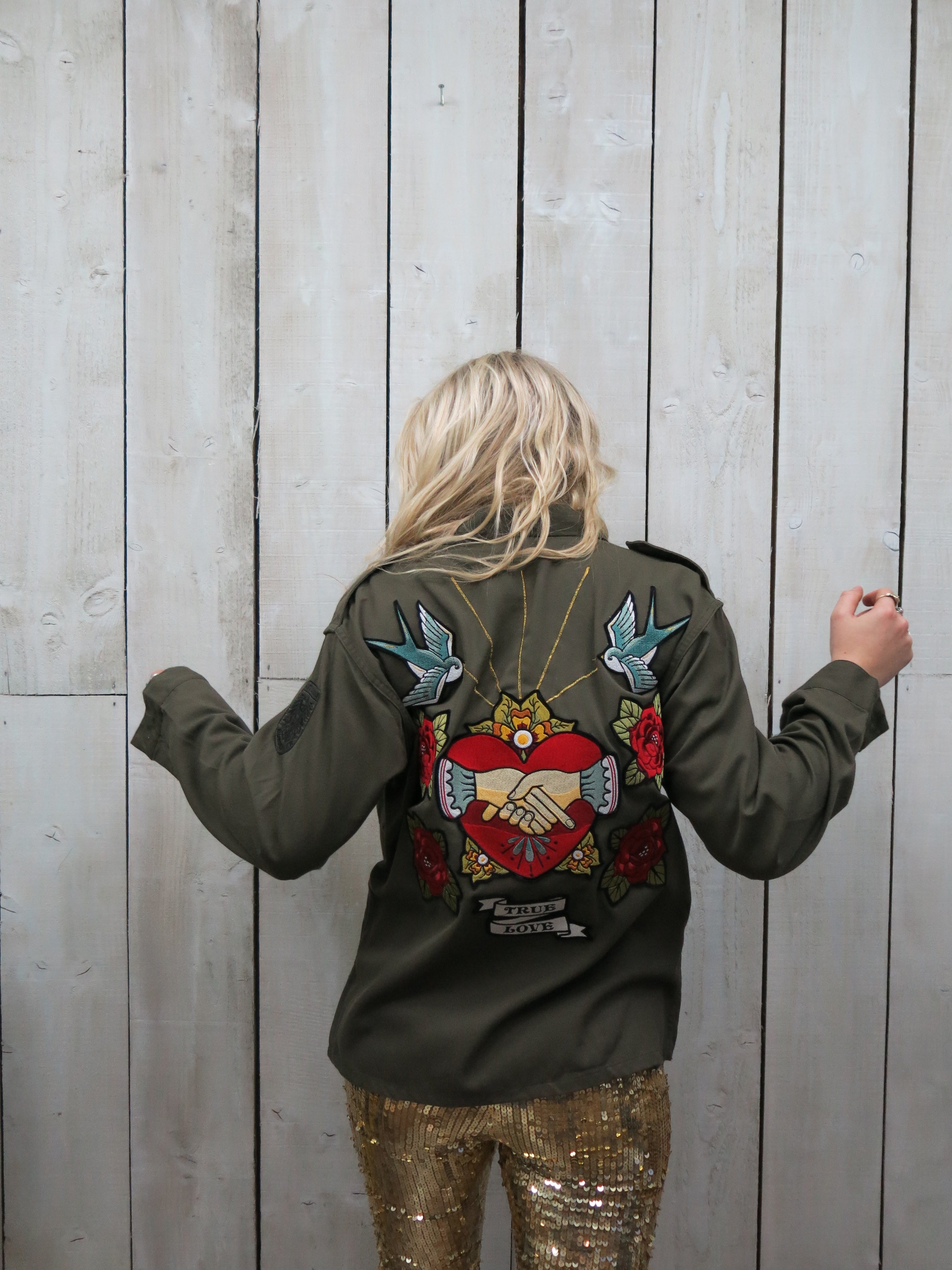 A woman viewed from behind wearing an olive green military jacket with a large embroidered back design featuring clasped hands inside a heart, swallows, roses, and the words "TRUE LOVE."