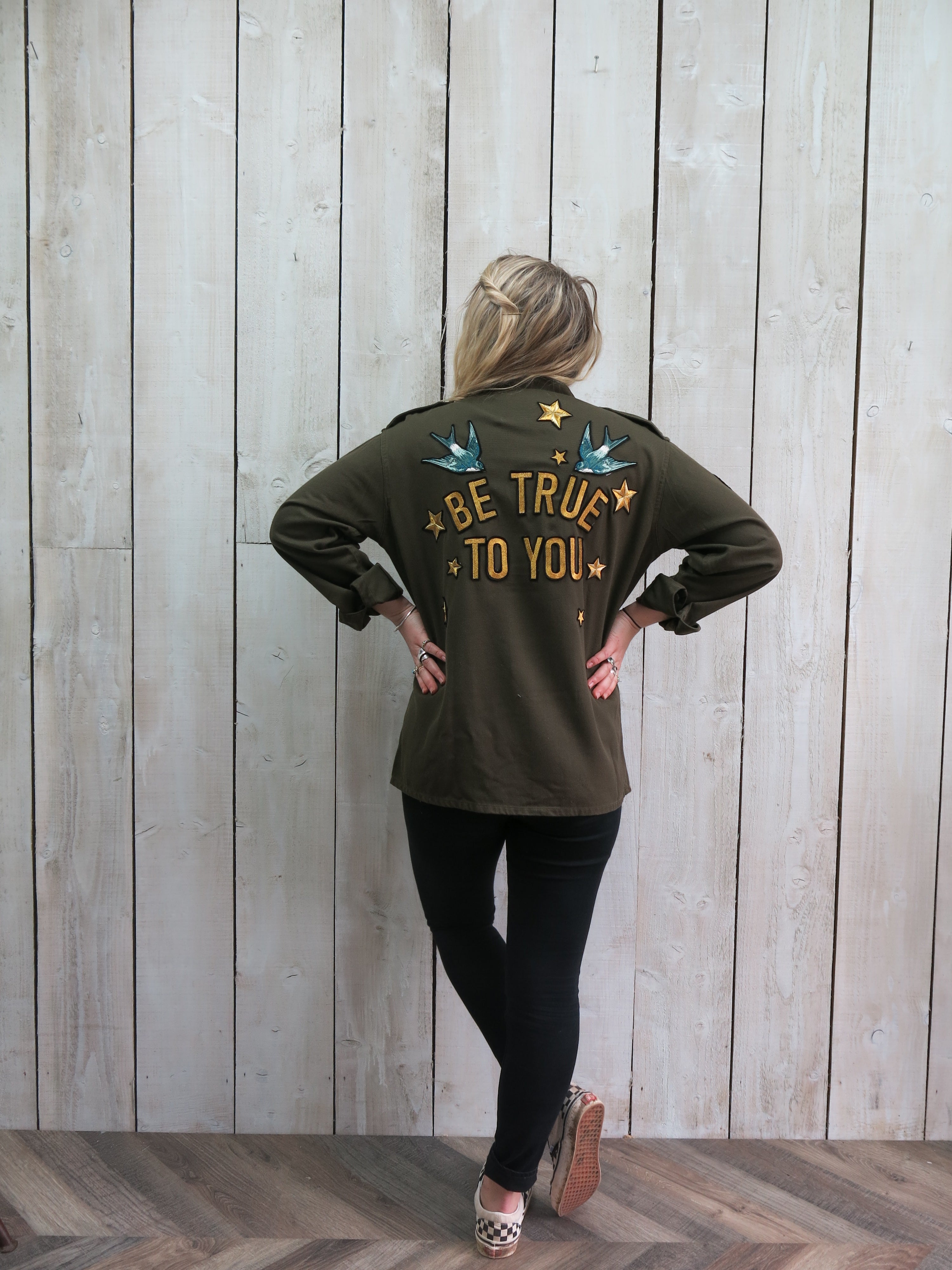Back view of a person wearing an olive green military jacket with "BE TRUE TO YOU" embroidered on the back, accented by blue birds and gold stars.