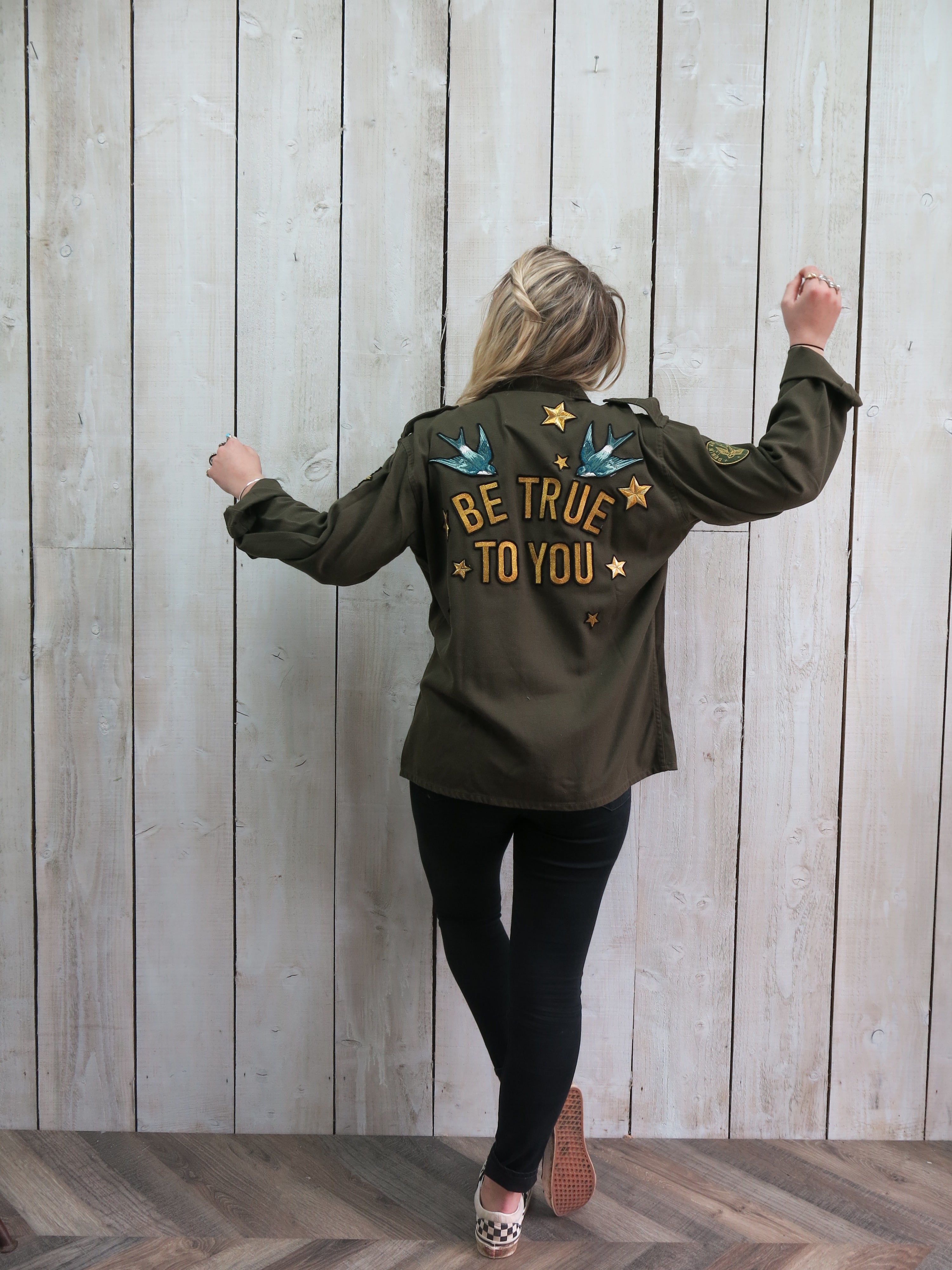 Back view of a person wearing an olive green military jacket with "BE TRUE TO YOU" embroidered on the back, accented by blue birds and gold stars.